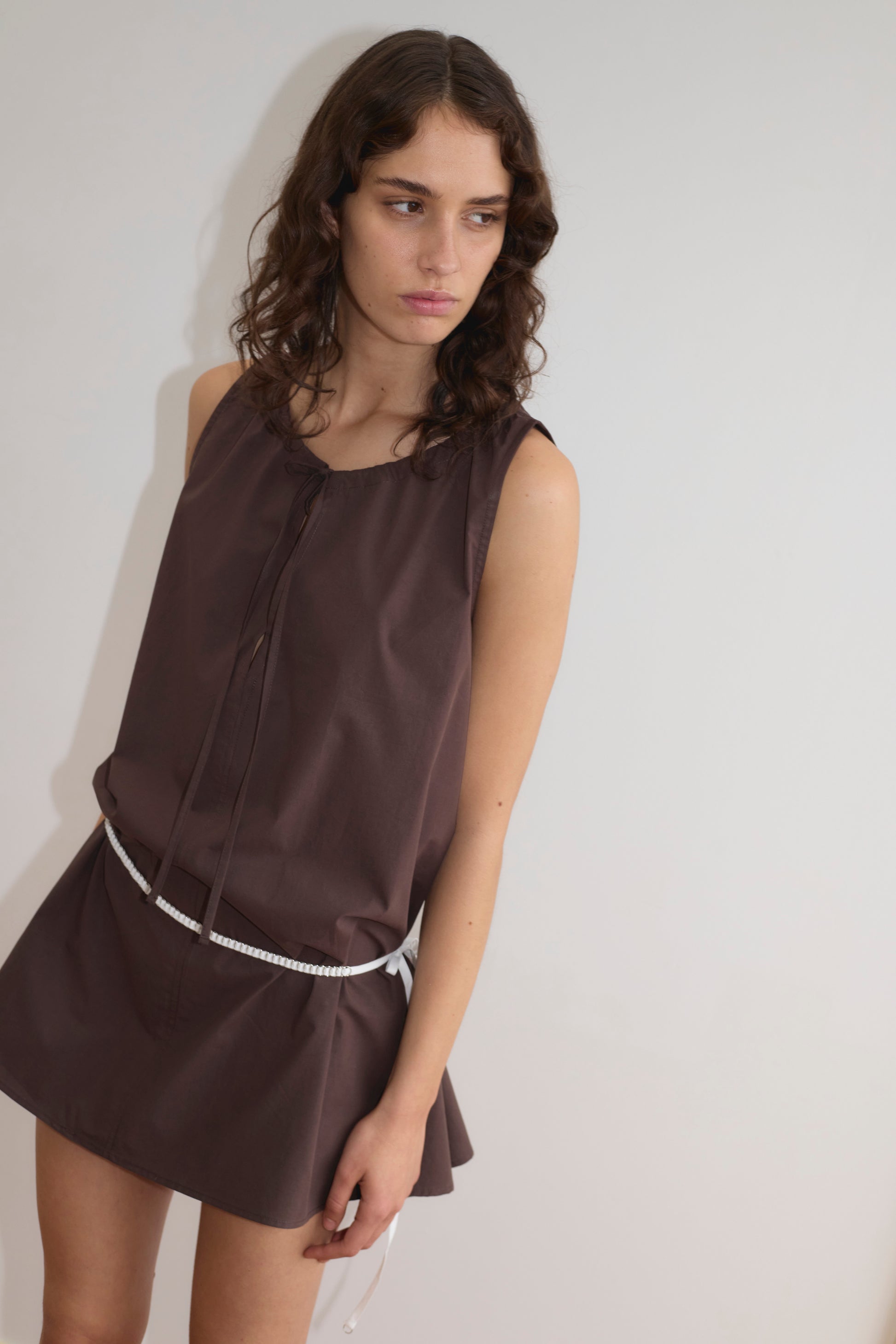 A person with wavy brown hair wears the sleeveless Drawcord Dress - Sepia by Deiji Studios, featuring an a-line silhouette and white belt, standing against a plain light wall and looking slightly left with a neutral expression.