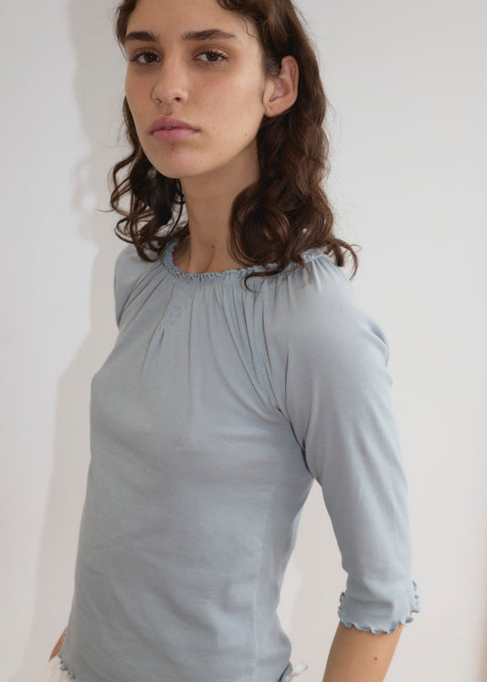 A person with wavy brown hair wears the Cotton Flounce 3/4 Sleeve - Baby Blue top, standing against a plain white background and gazing slightly to the side with a neutral expression.