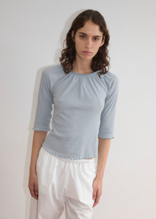 A person with wavy brown hair wears Deiji Studios’ Cotton Flounce 3/4 Sleeve - Baby Blue top and loose white pants, standing against a plain white background with a neutral expression.