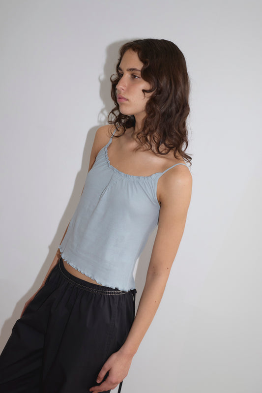 A person with long, wavy brown hair wears the Cotton Flounce Tank - Baby Blue from Deiji Studios and black pants, standing against a plain white background and looking slightly to the side with a neutral expression.