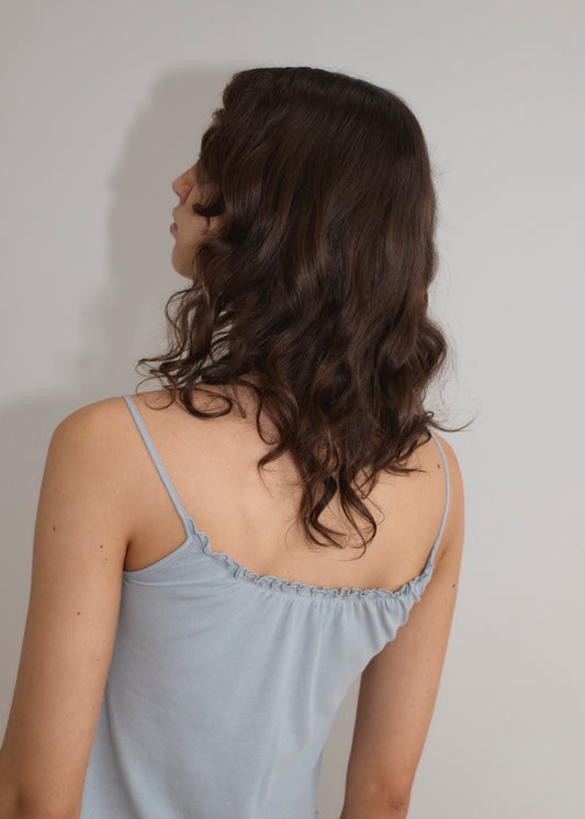 A woman with wavy brown hair faces away from the camera, wearing the Cotton Flounce Tank in Baby Blue by Deiji Studios, against a plain light-colored wall.