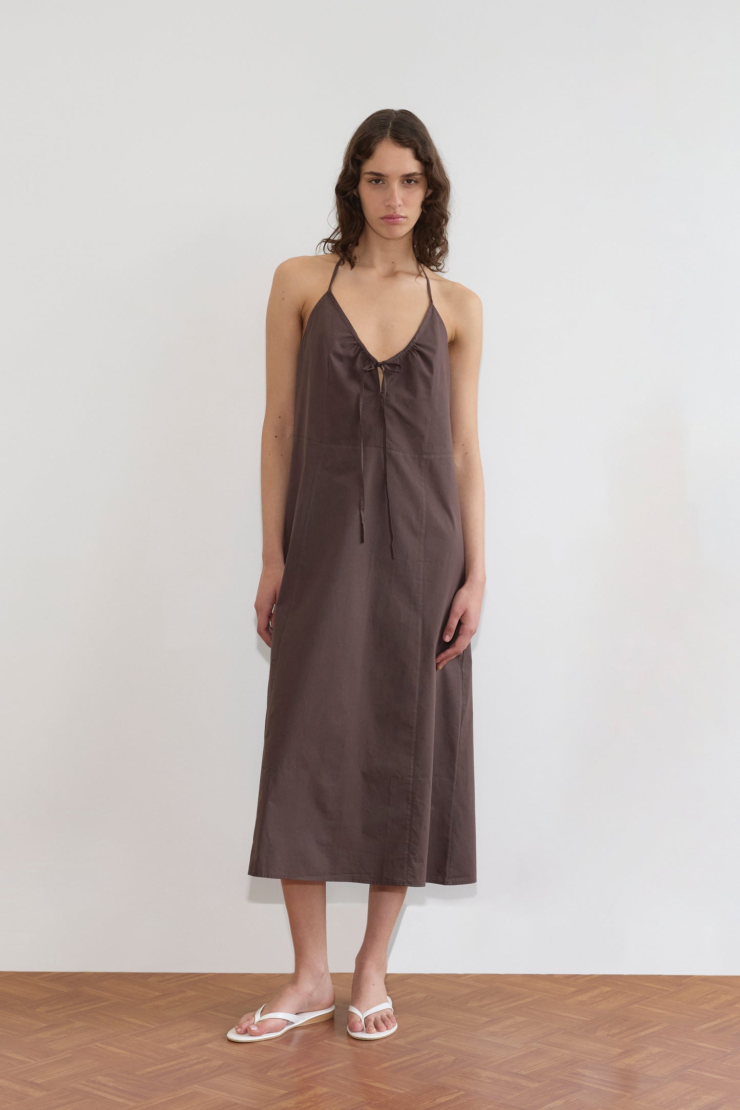A woman with wavy brown hair wears the Seamed Halter Dress - Sepia by Deiji Studios, featuring a sleeveless halter cut and front tie detail. She stands on a wooden floor against a white wall, styled with white flip-flops.