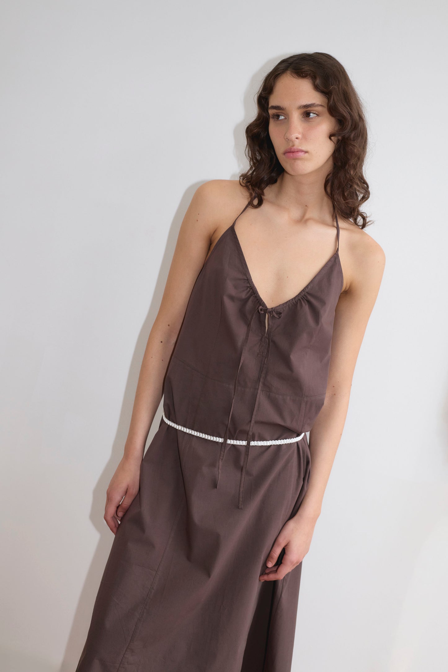 A woman with wavy brown hair wears the Seamed Halter Dress, a sleeveless deep V-neck brown dress with a thin white belt, standing against a plain white background.