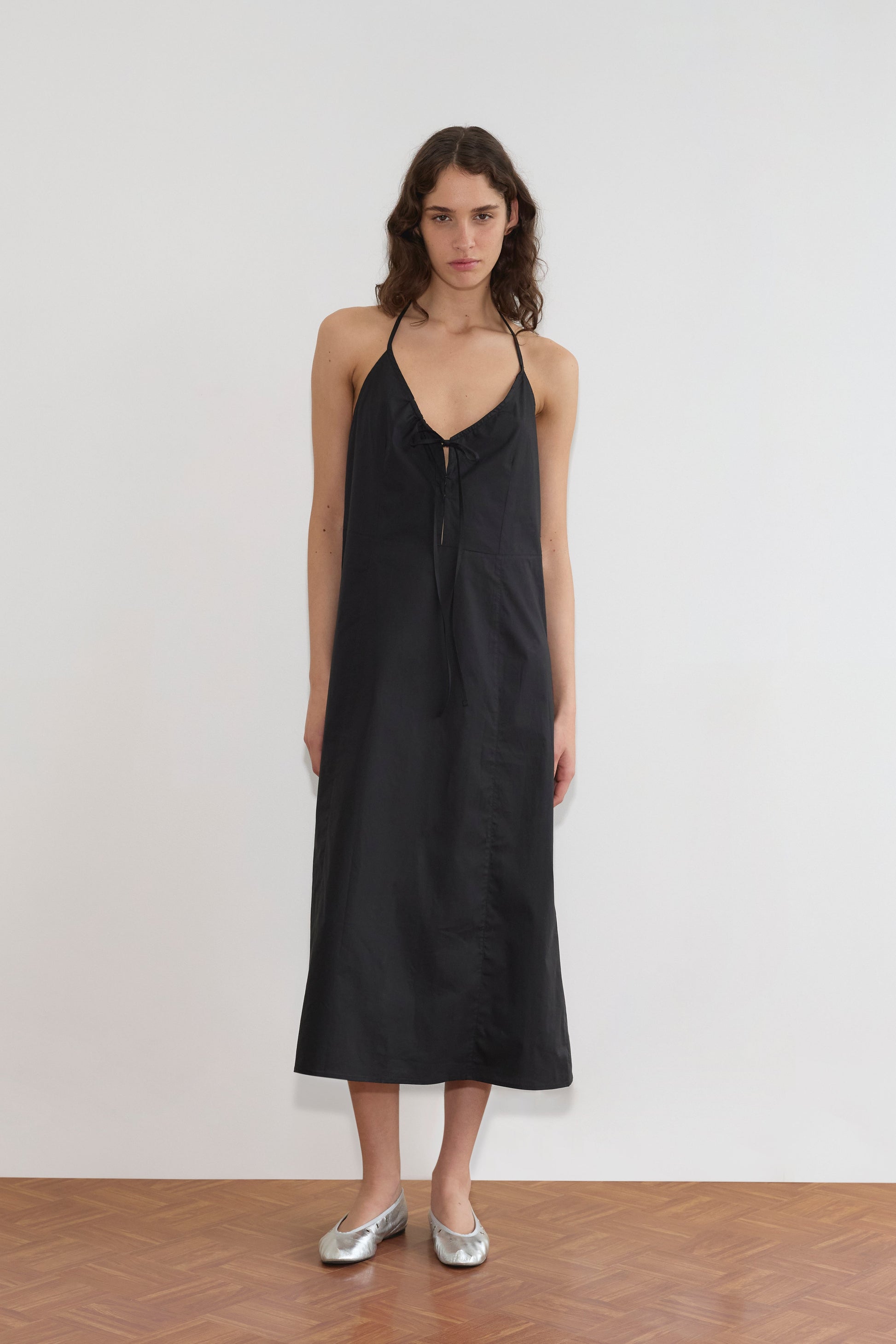 A woman with long brown hair wears the Seamed Halter Dress - Black, styled with silver ballet flats. She stands indoors on a wooden floor against a plain white wall.