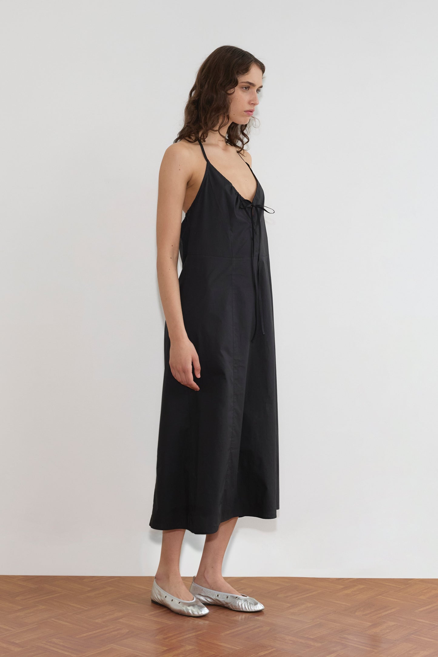 A woman stands sideways against a plain white wall, wearing the Seamed Halter Dress - Black with thin straps and bow detail, paired with silver flat shoes on a wooden floor.