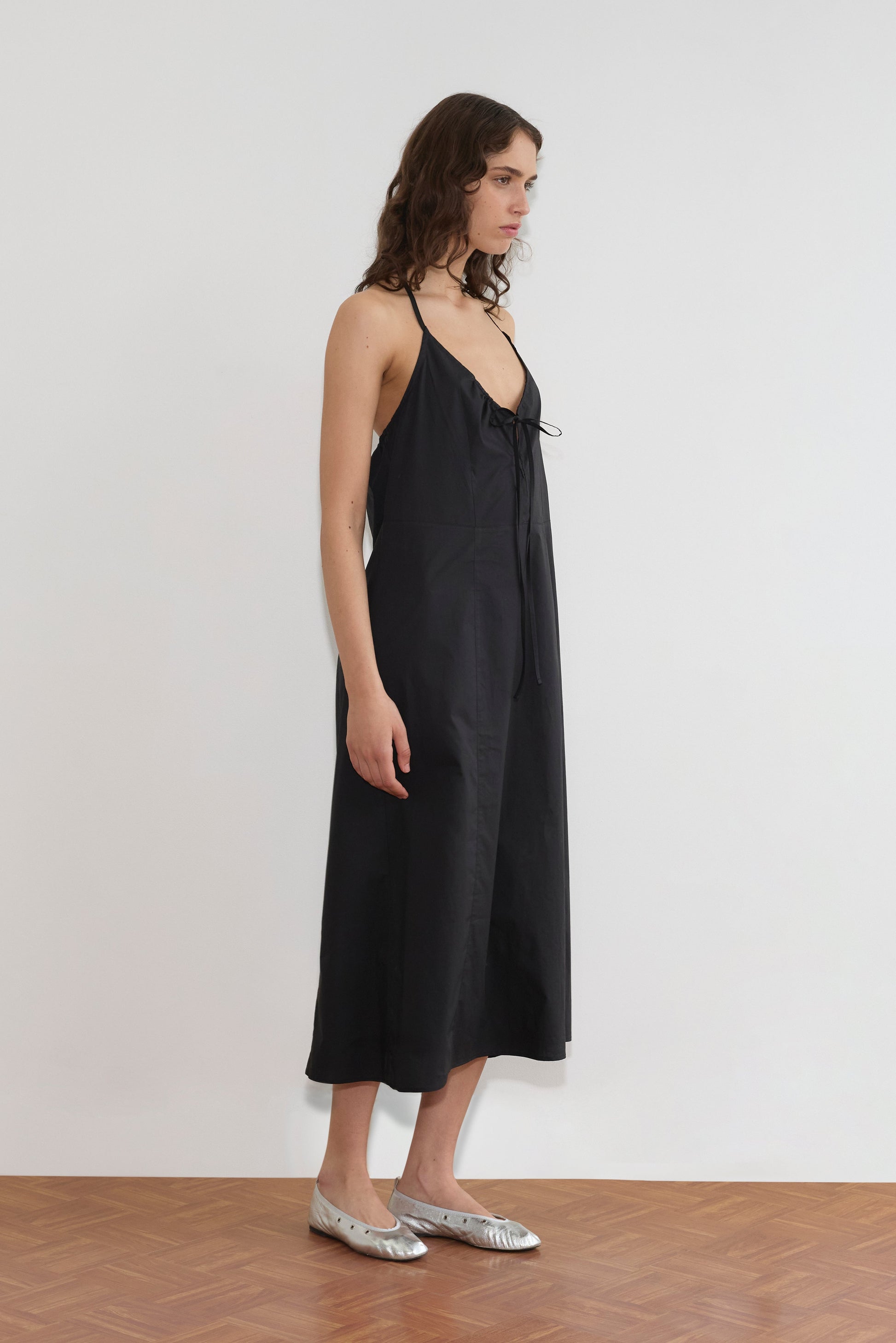 A woman stands sideways against a plain white wall, wearing the Seamed Halter Dress - Black with thin straps and bow detail, paired with silver flat shoes on a wooden floor.