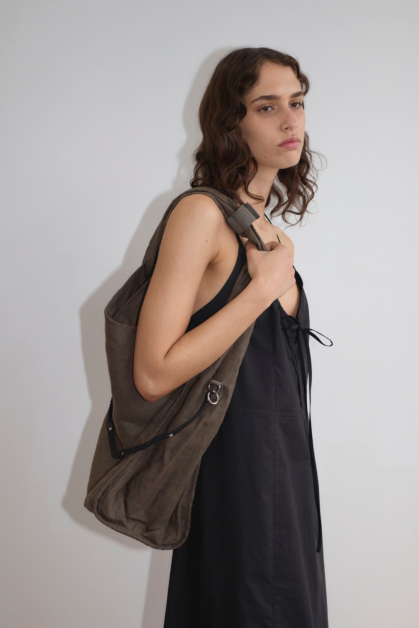 A person with wavy brown hair wearing the Seamed Halter Dress - Black stands against a plain white wall, holding a large brown shoulder bag from Deiji Studios.