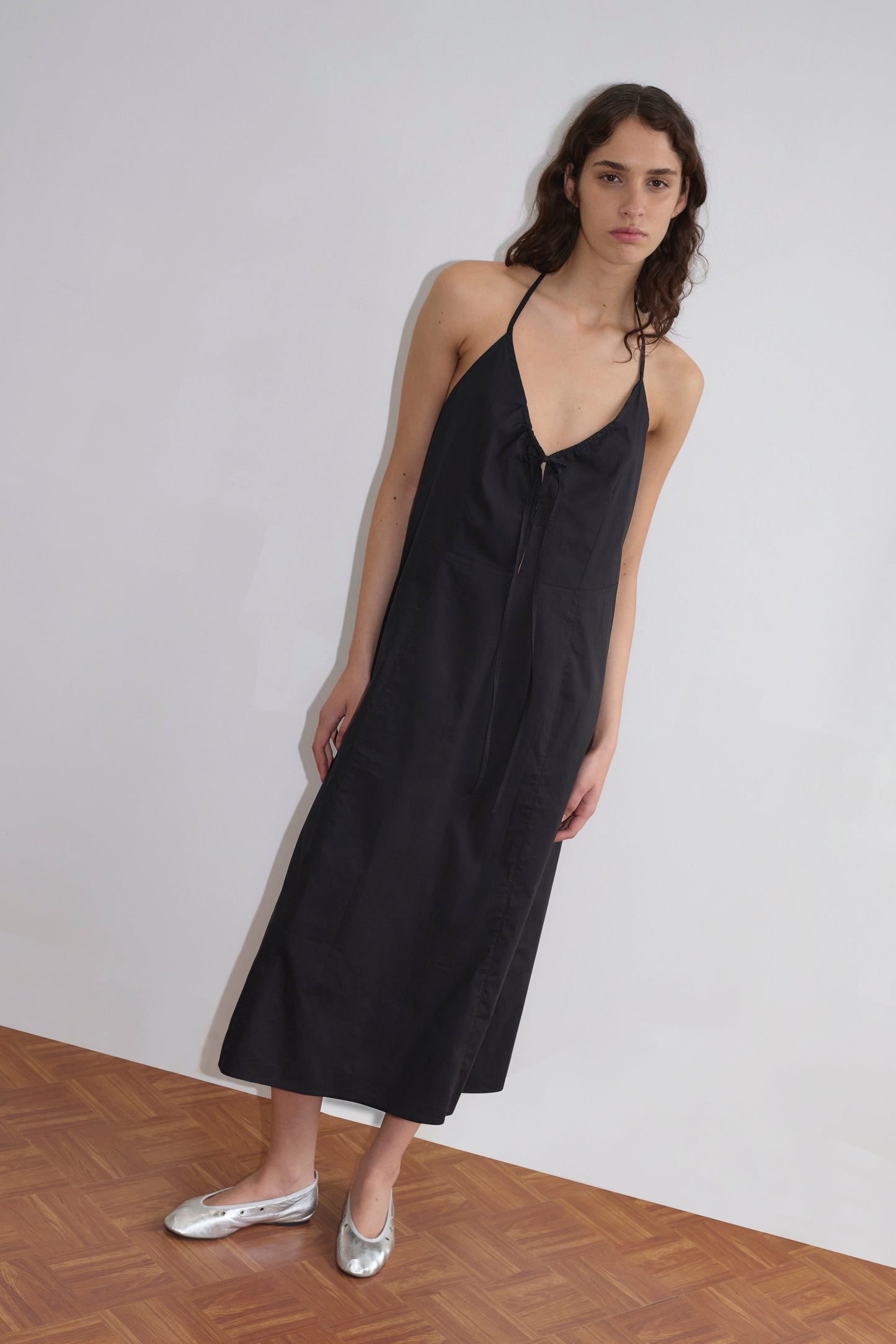 A person with long, wavy brown hair wears the black Seamed Halter Dress and metallic silver ballet flats, standing on a wooden floor against a plain white wall.