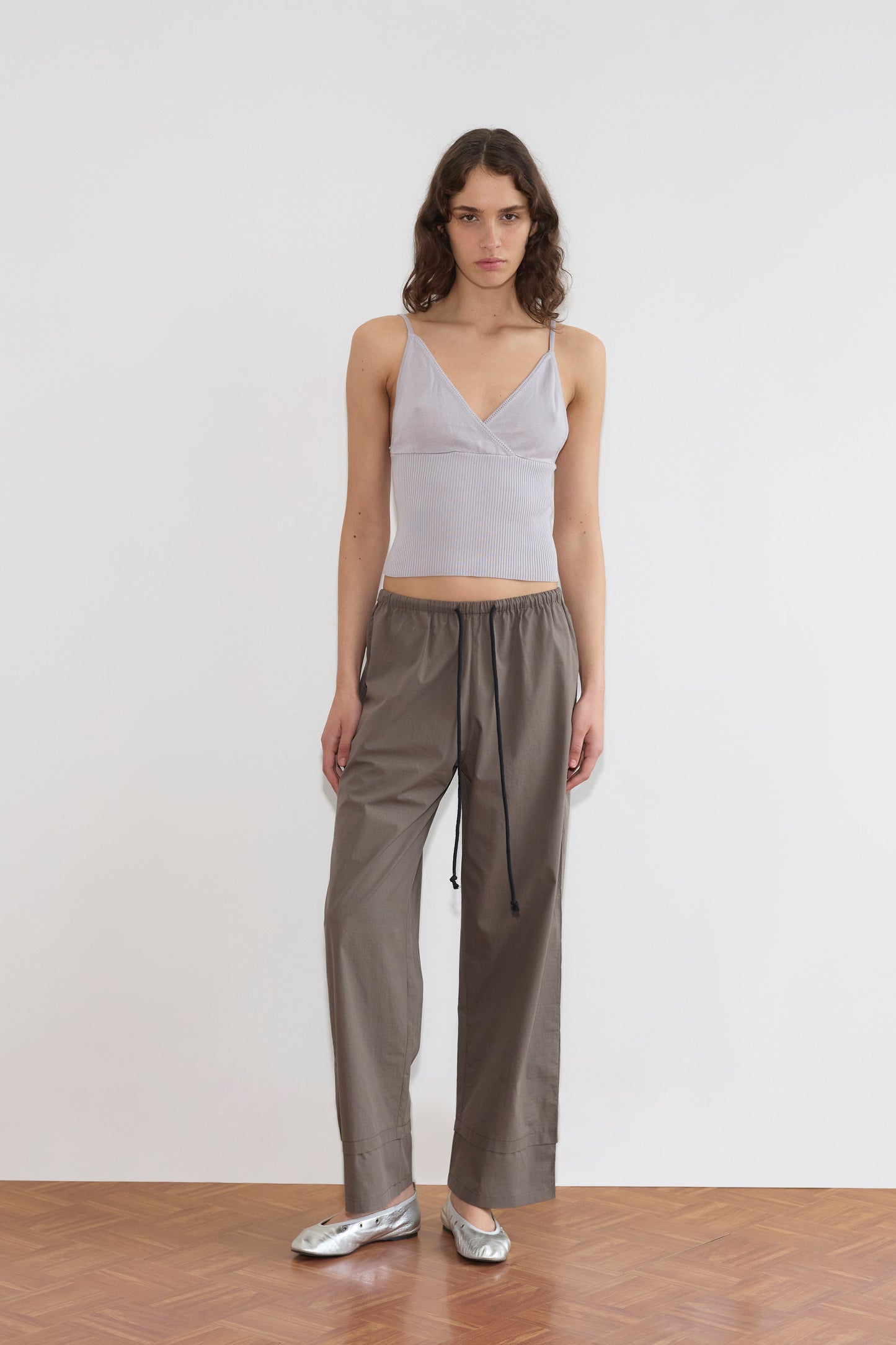 Model wears the Cross Panel Knit Top in soft lilac with olive grey wide-leg drawstring trousers and silver ballet flats, standing against a white wall for an effortless Deiji Studios look.