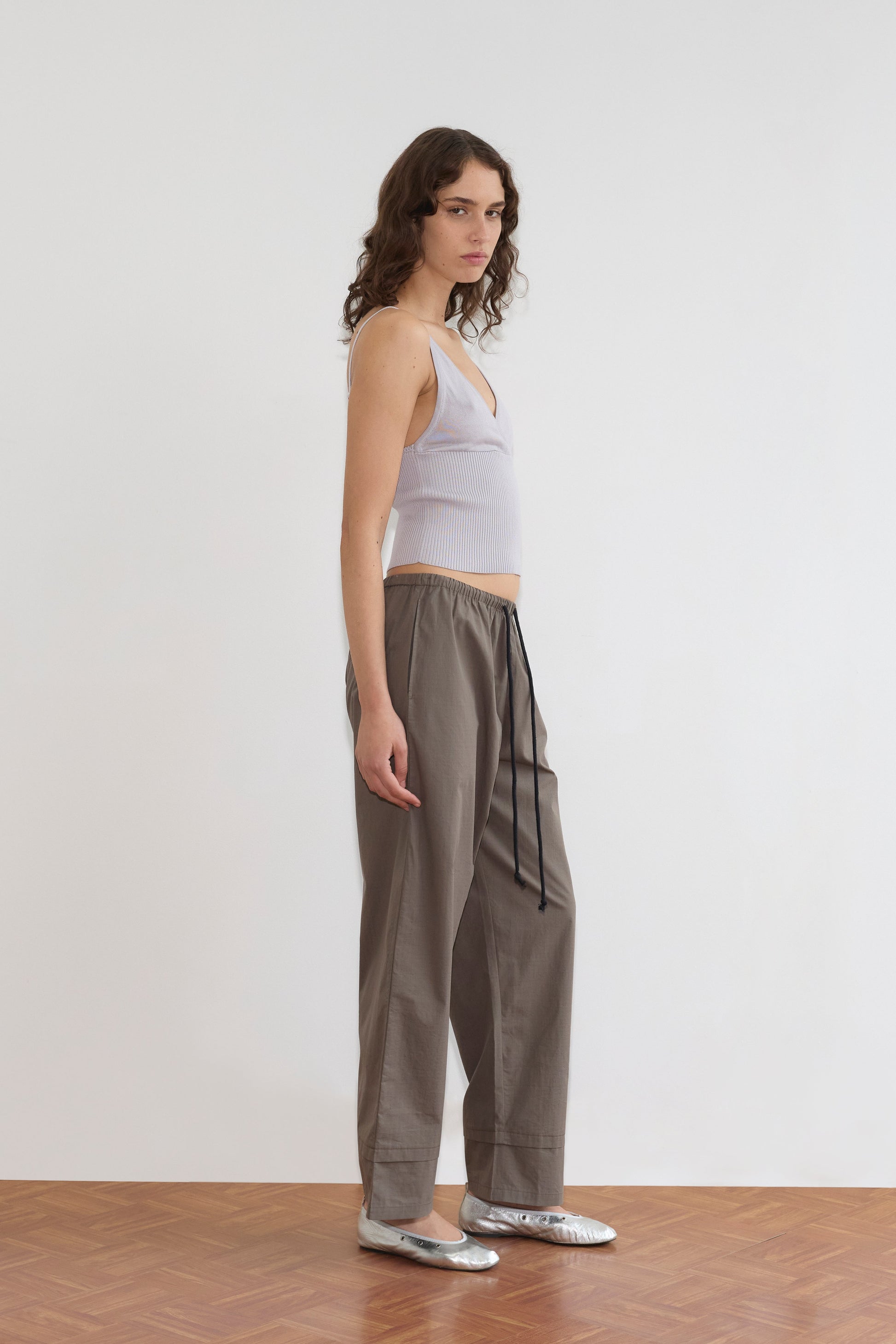 A woman with wavy brown hair stands sideways on a wooden floor, wearing the Cross Panel Knit Top in Soft Lilac, loose gray drawstring pants from Deiji Studios, and silver flats against a plain white background.