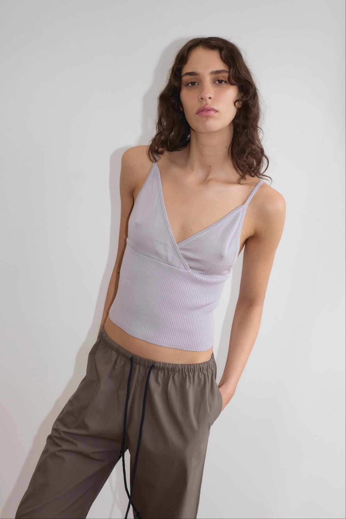A person with long, wavy brown hair wears a Cross Panel Knit Top in light purple and loose brown drawstring pants, standing against a plain white wall with a relaxed expression.