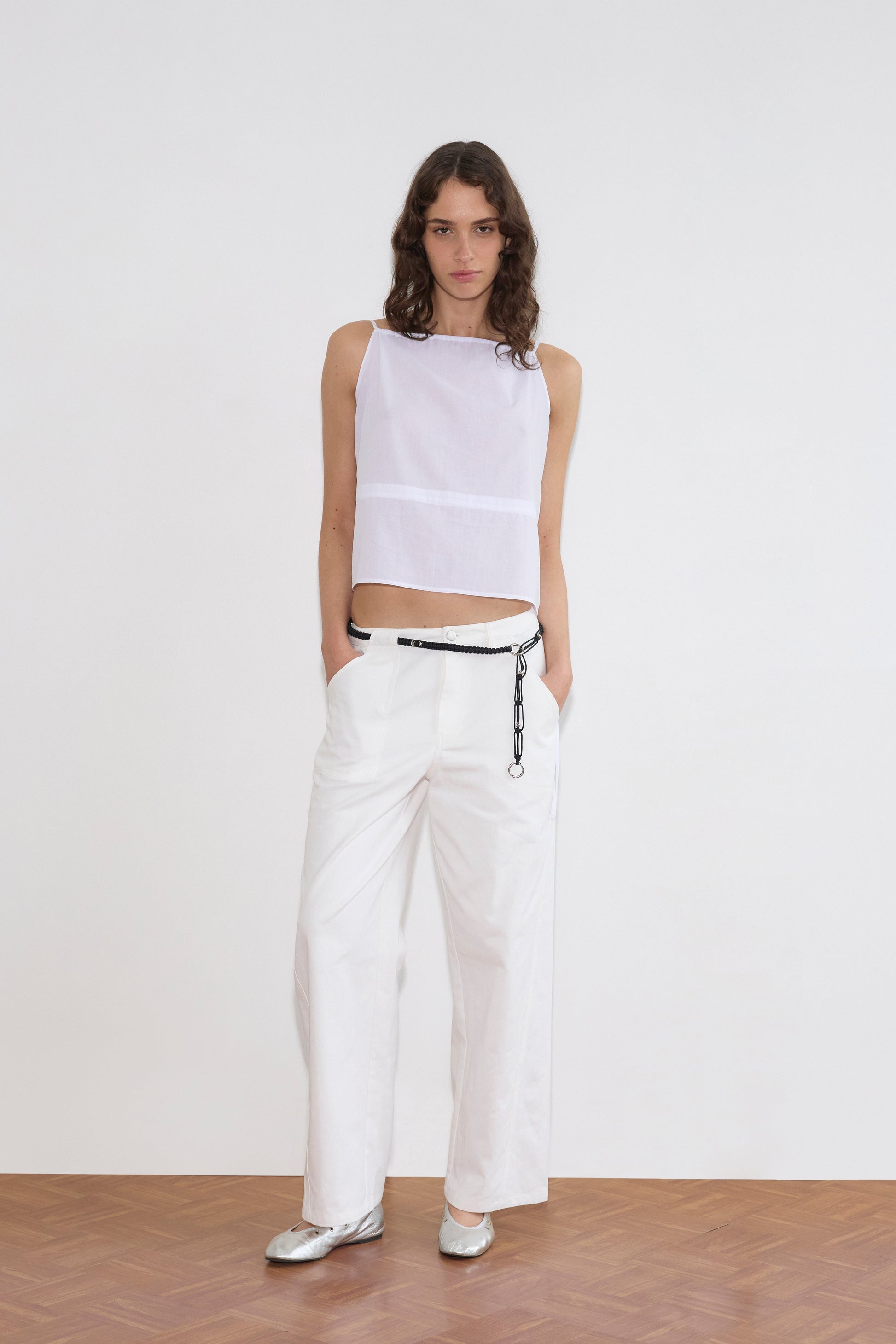 A person stands against a plain white background wearing a sleeveless white crop top, the Pocket Jean - White styled low-rise with hands in pockets, a thin black belt with a chain, and silver ballet flats.