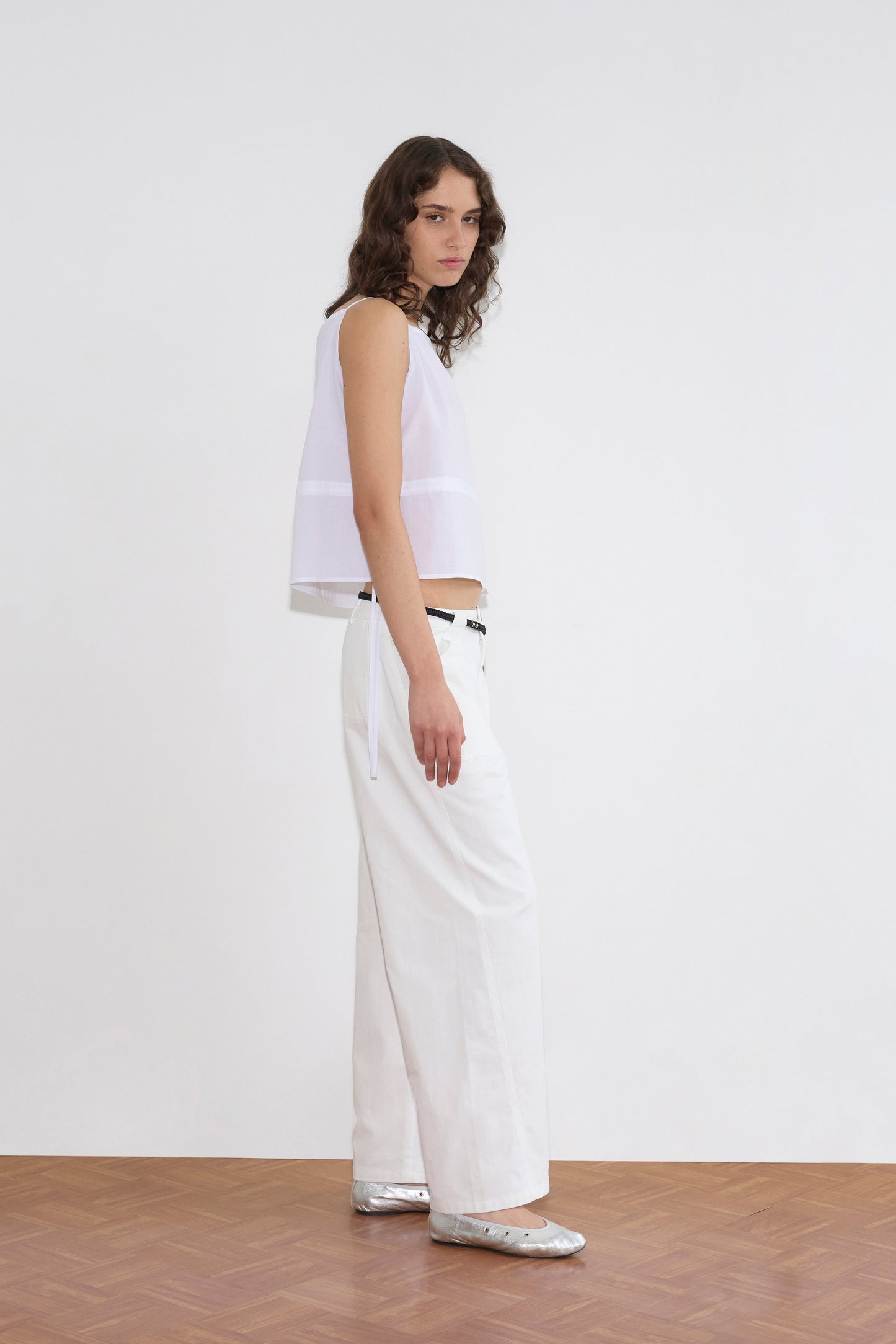 A person with wavy brown hair stands sideways on a wooden floor, wearing a sleeveless white top and the Pocket Jean - White, paired with a black belt and silver shoes against a plain white background.