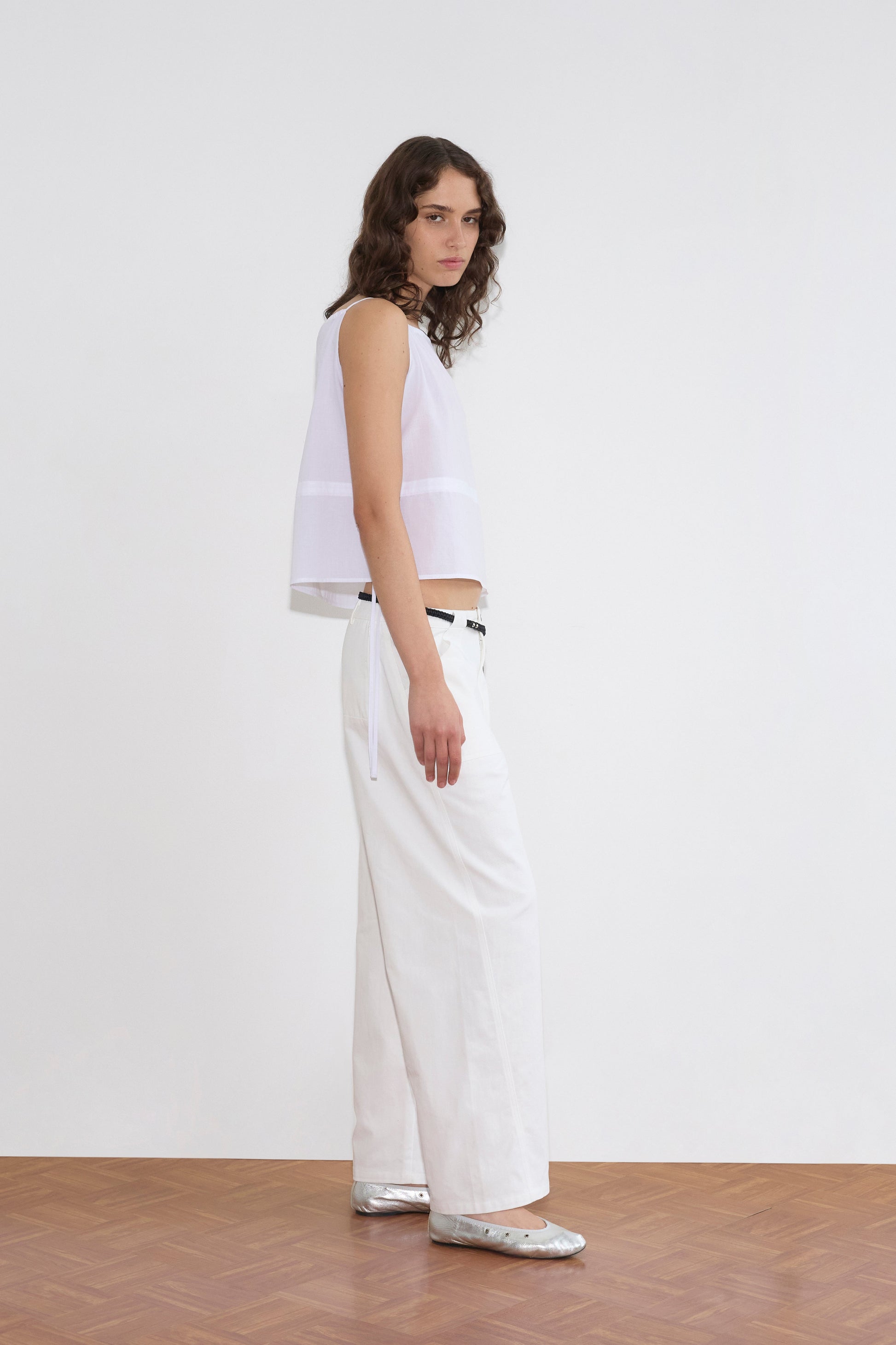 A person with wavy brown hair stands sideways on a wooden floor, wearing a sleeveless white top and the Pocket Jean - White, paired with a black belt and silver shoes against a plain white background.