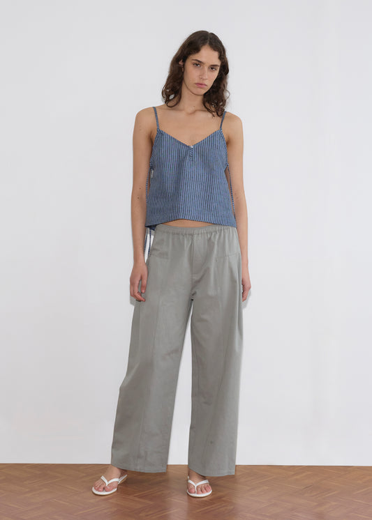 A woman with long, wavy brown hair and a neutral expression stands on a wooden floor against a white background, wearing the V Neck Cami - Azure Stripe, loose light gray pants, and white sandals.