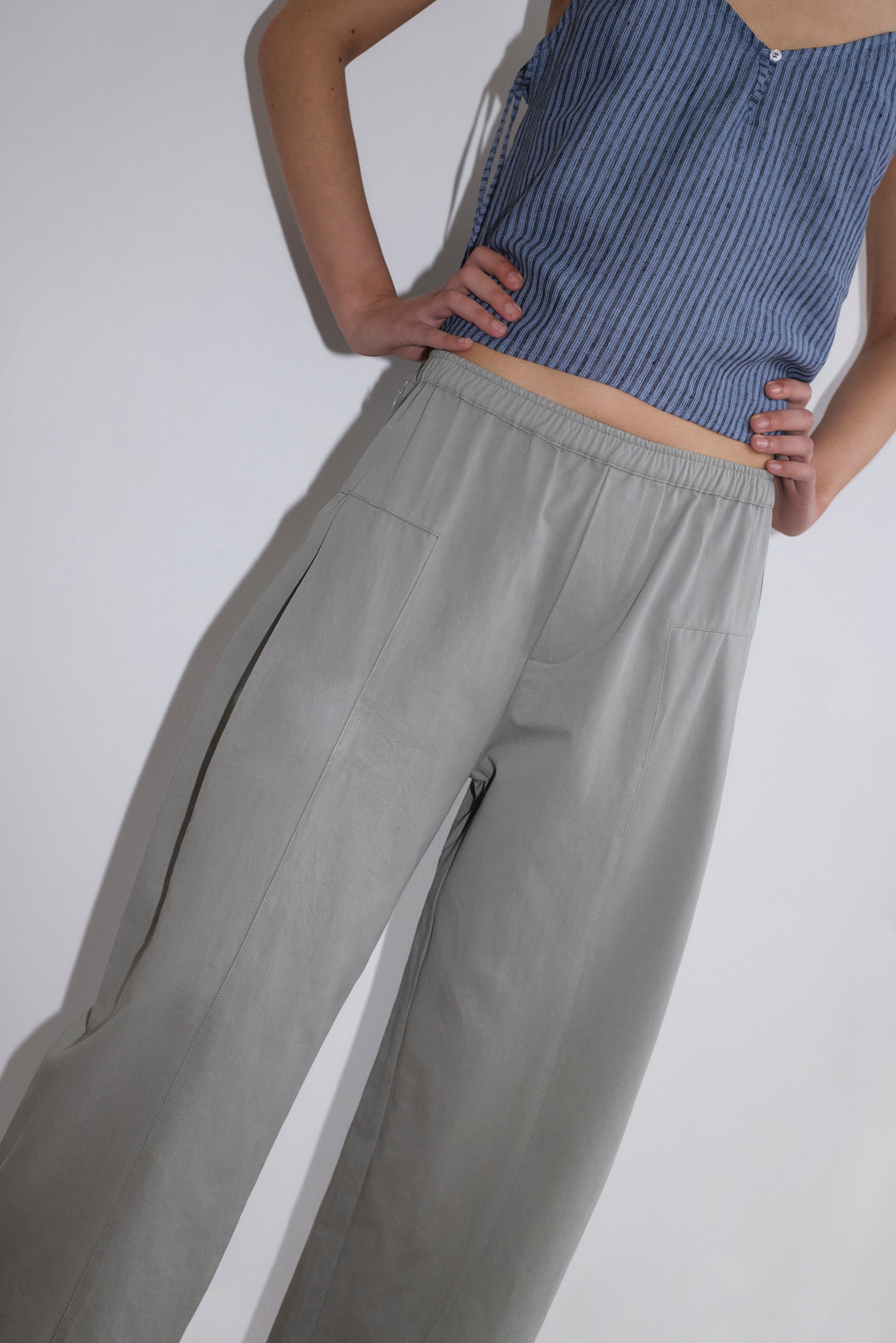 A person wears the Deiji Studios V Neck Cami - Azure Stripe with loose light gray pants, standing hands on hips against a plain white background. The image is cropped at the shoulders and mid-calf.