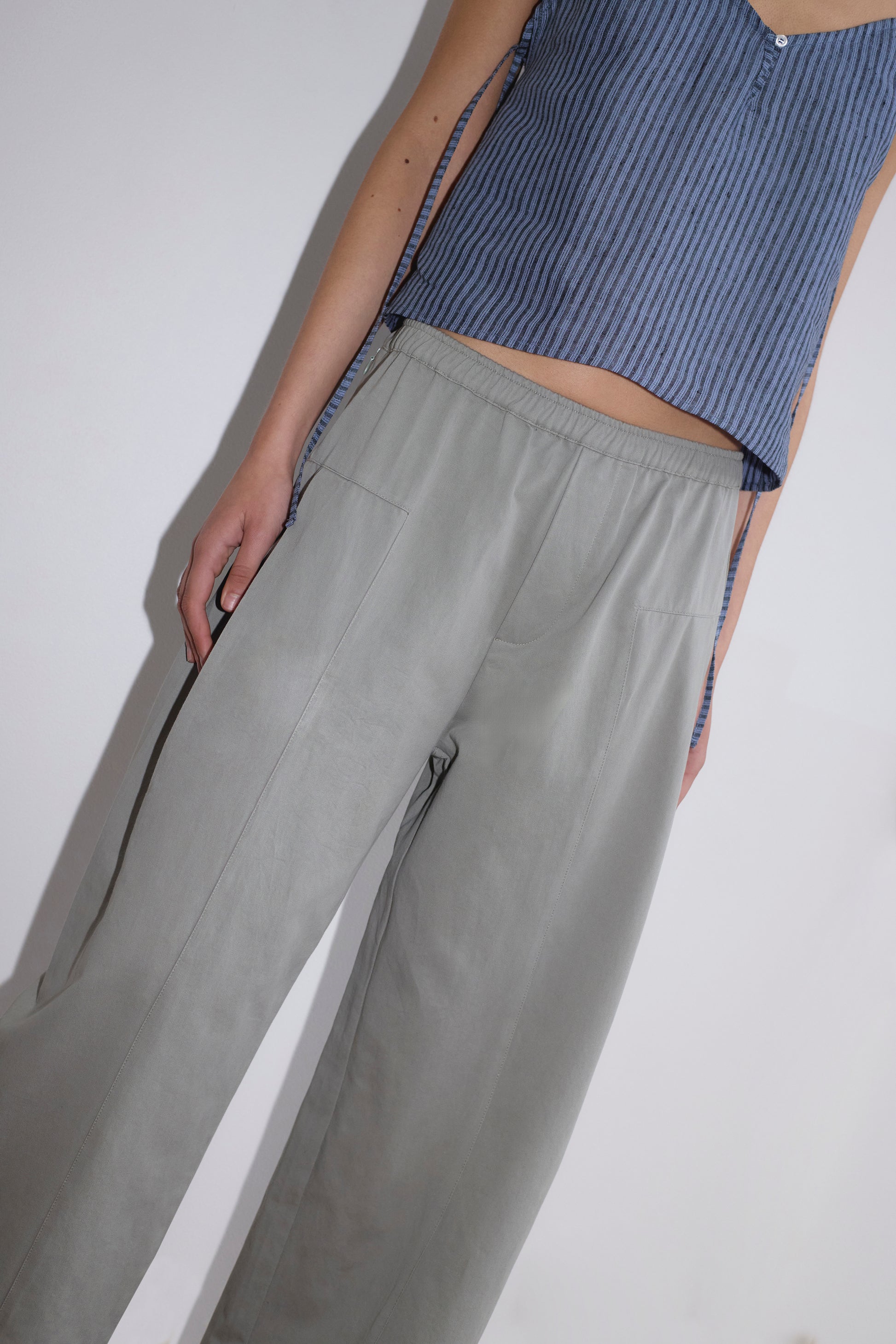 A person models the Pleat Pant - Sage Grey, featuring a relaxed straight leg, paired with a sleeveless blue pinstriped top, shown from the shoulders down against a plain background.