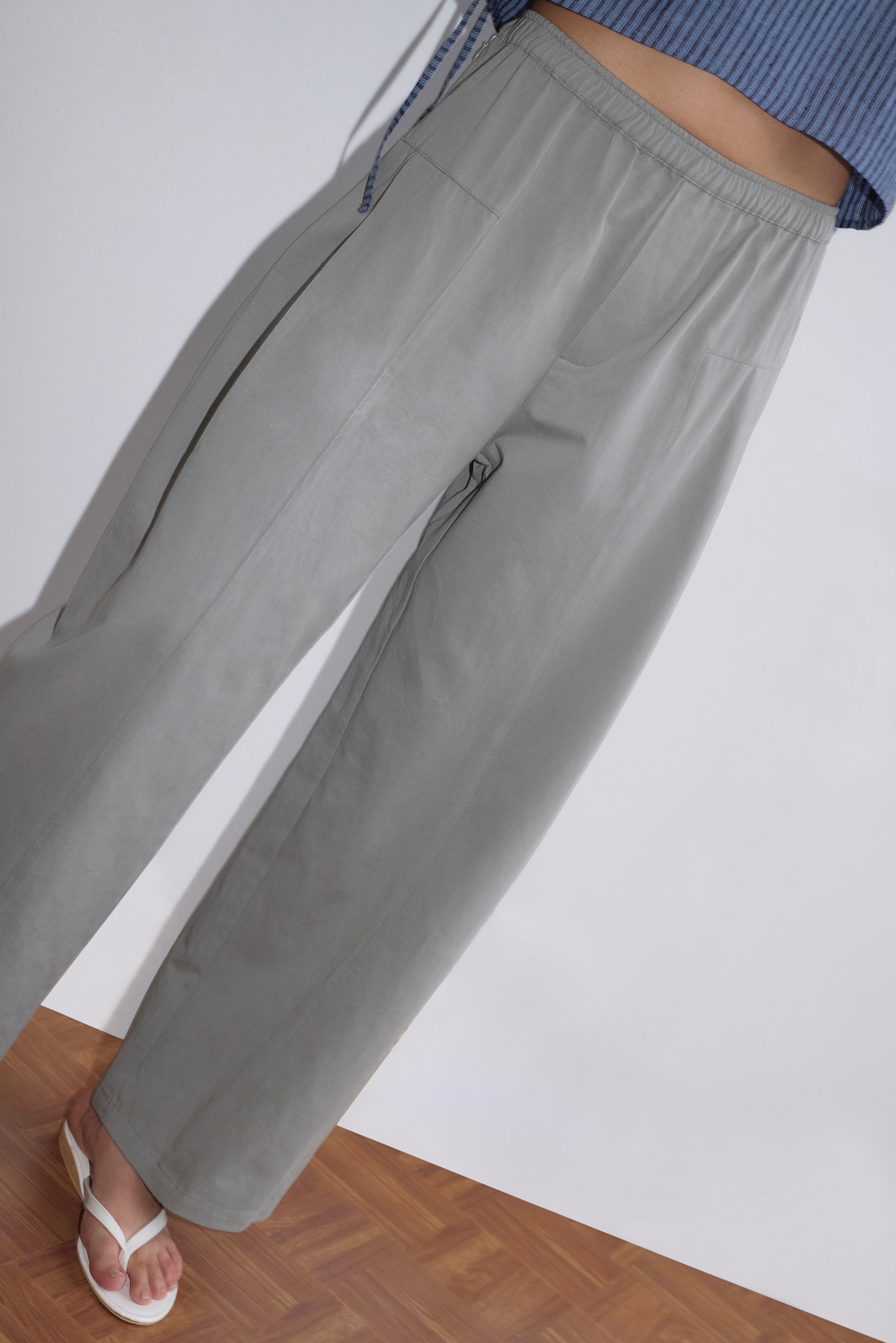 A person wearing the Pleat Pant - Sage Grey, which is high-waisted with a relaxed straight leg and elastic waistband, paired with a blue ribbed top and white sandals, stands on a wood floor against a white background.