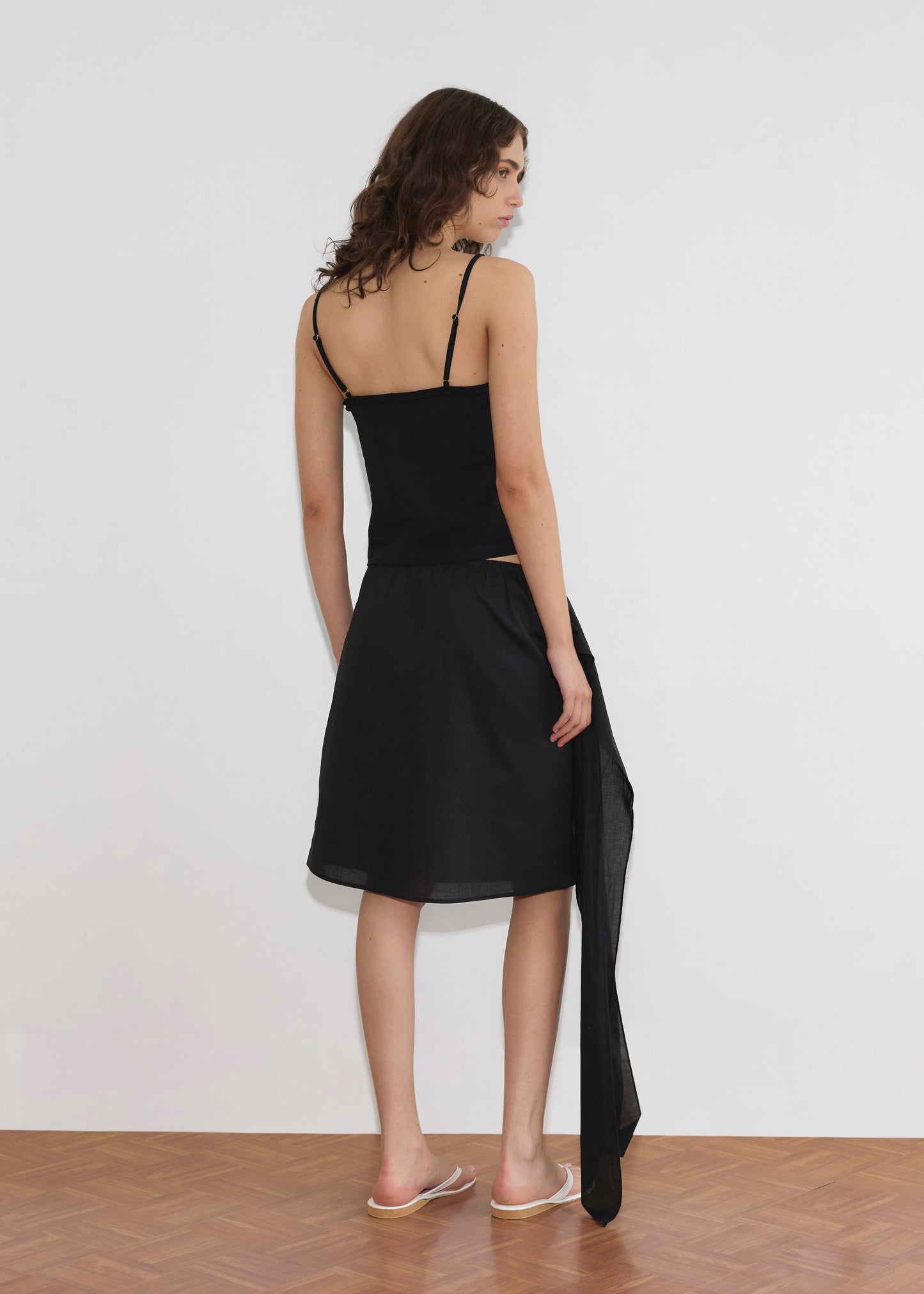 A woman with wavy brown hair stands facing a white wall, wearing a black sleeveless top, the Scarf Midi - Black by Deiji Studios featuring an asymmetrical sheer overlay, and clear slide sandals on a wooden floor.