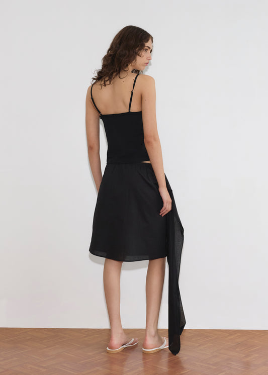 A woman with wavy brown hair stands facing a white wall, wearing a black sleeveless top, the Scarf Midi - Black by Deiji Studios featuring an asymmetrical sheer overlay, and clear slide sandals on a wooden floor.