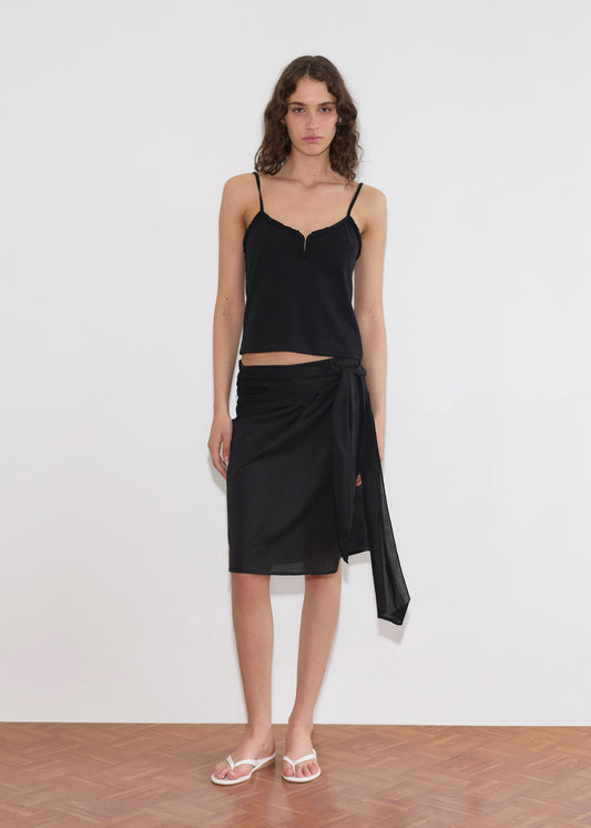 A woman with wavy brown hair wears the Clasp Singlet - Black by Deiji Studios, paired with a black wrap skirt and white flip-flops. She stands on a wooden floor against a plain white wall.
