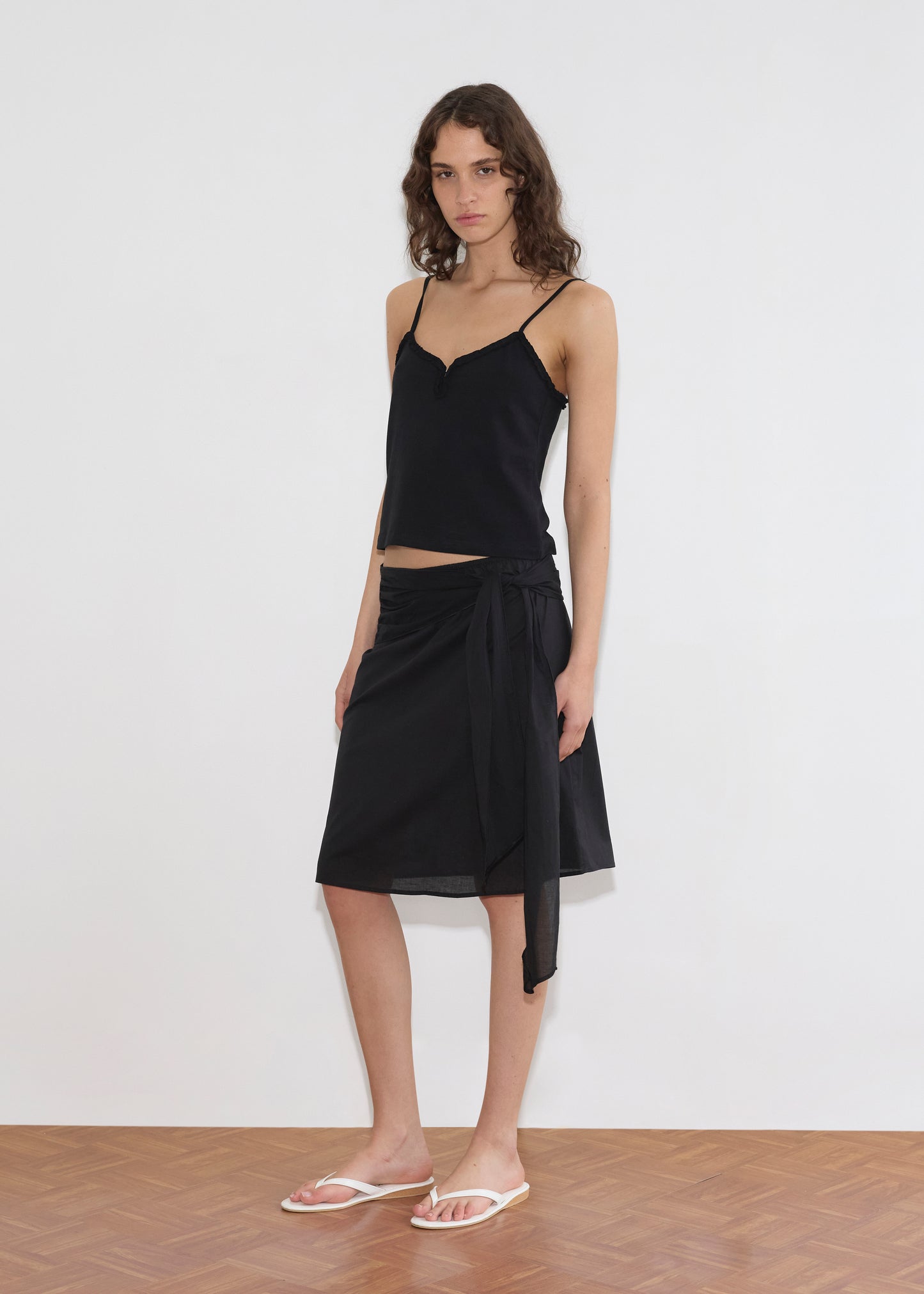 A woman with wavy hair and a neutral expression stands against a plain white wall, wearing the Deiji Studios Scarf Midi - Black dress and white flip-flops.