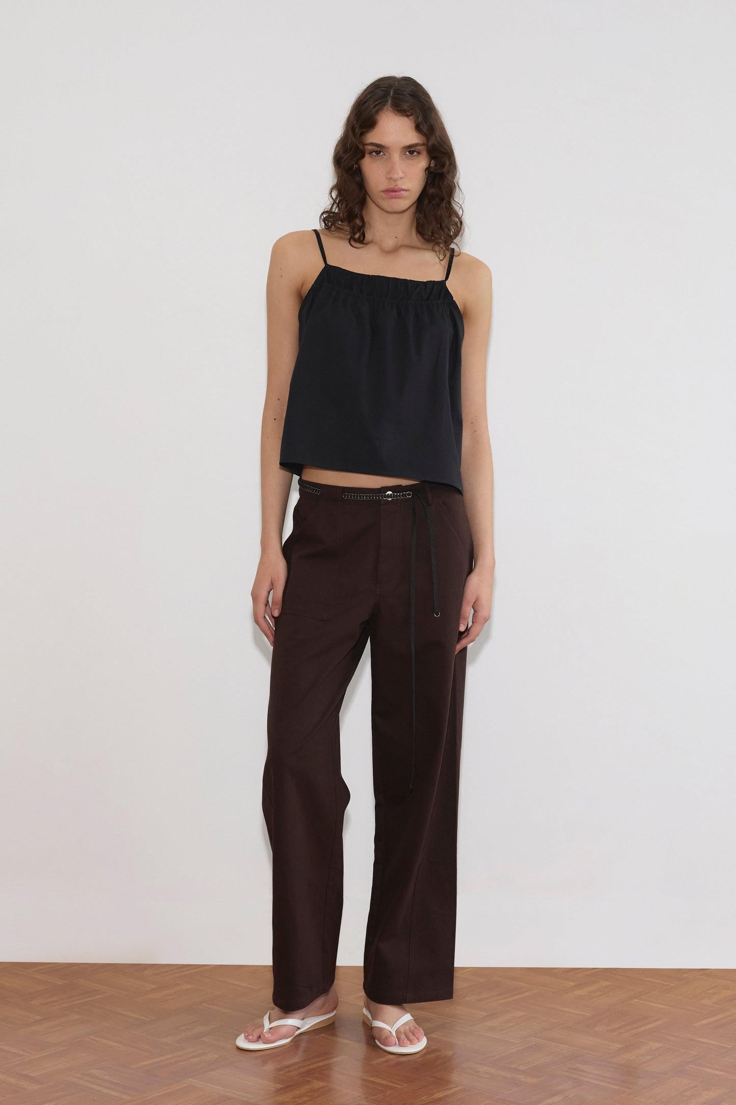 A person with wavy hair wears a loose black camisole top, Deiji Studios Pocket Jean in dark brown with a thin belt, and clear sandals, standing on a wooden floor against a plain white background.