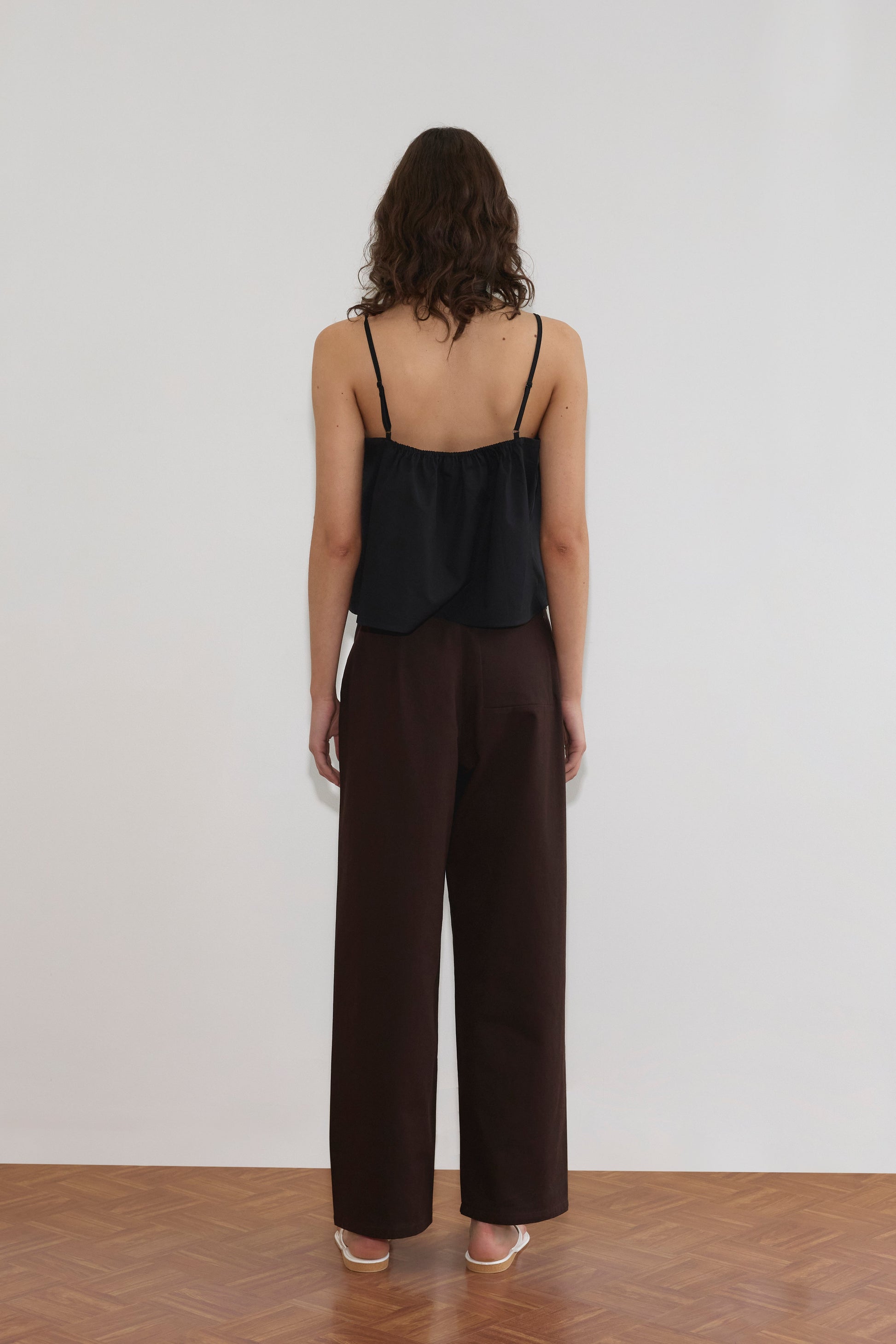 A person with wavy brown hair stands facing a white wall, wearing a black sleeveless top, Pocket Jean - Dark Brown by Deiji Studios, and white heeled sandals on a wooden floor.