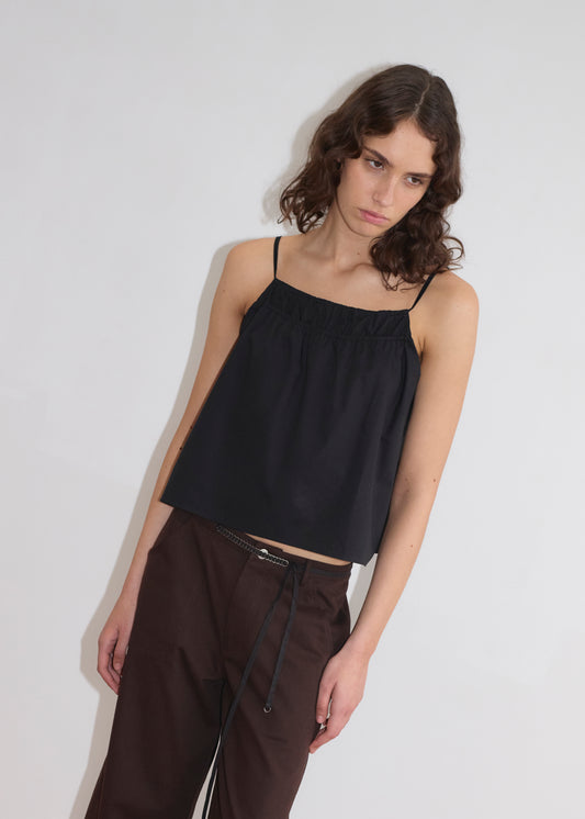 A woman with wavy brown hair wears the Double Layer Top - Black by Deiji Studios and dark pants, standing against a plain white background, looking slightly downward with a neutral expression.