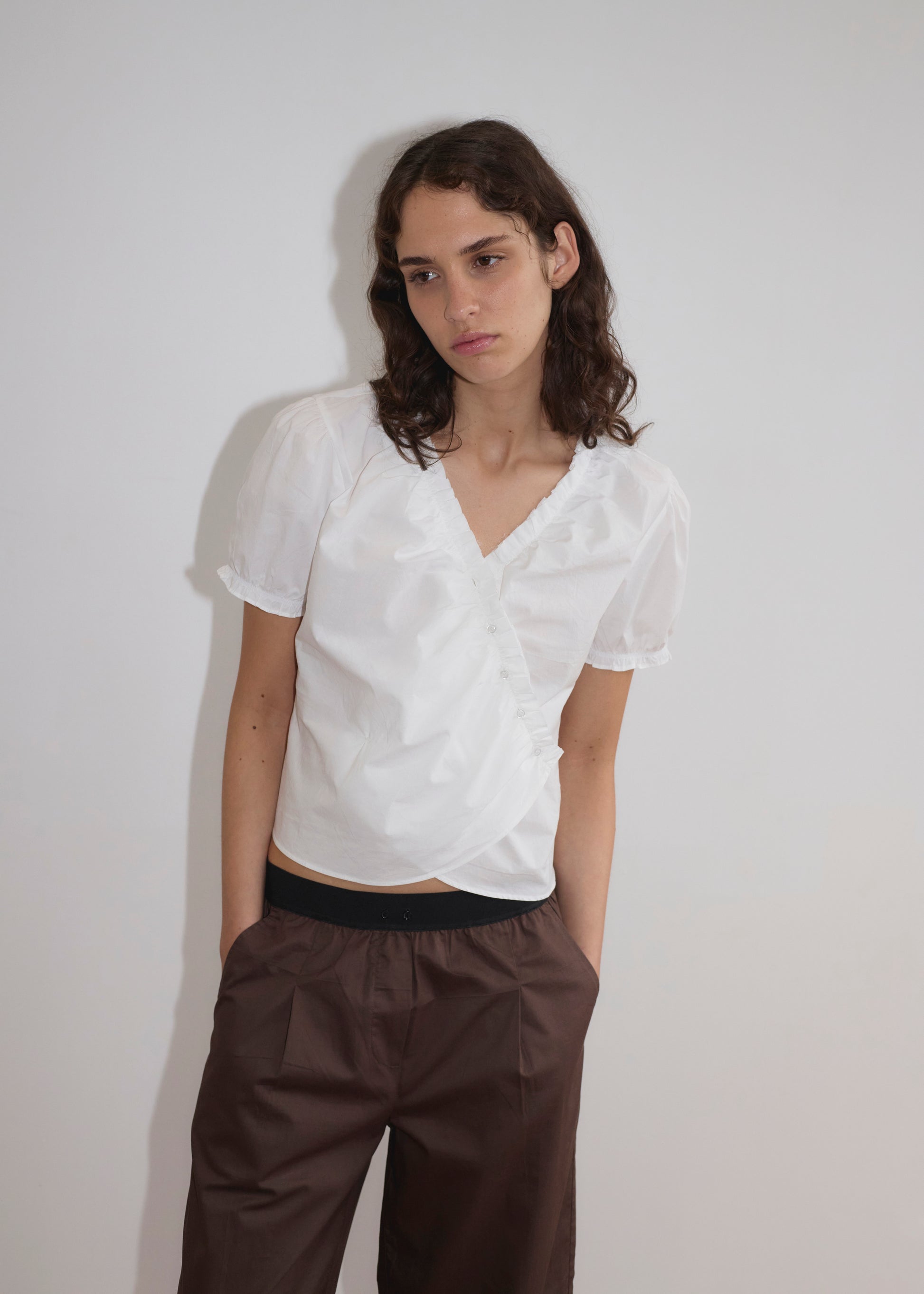A person with long brown hair wears a white short-sleeve blouse and the Contrast Waist Pant - Brown by Deiji Studios in a relaxed straight leg, standing against a plain white wall with hands in pockets, looking slightly to the side neutrally.