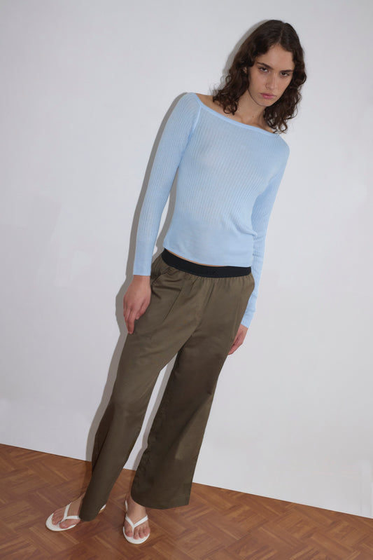 A person with curly hair wears a light blue long-sleeve top, the Contrast Waist Pant - Olive by Deiji Studios in a relaxed straight leg fit, and white flip-flops, standing against a plain white background on a wooden floor.