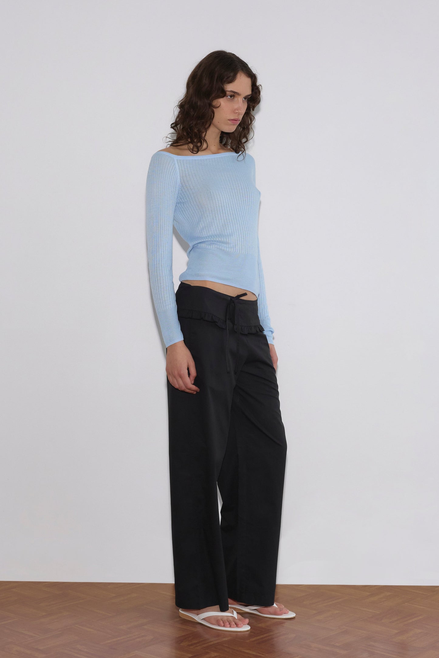 A person with wavy brown hair stands against a white wall on a wooden floor, wearing a light blue off-shoulder ribbed top, Flounce Belt Pant in black, and white flip-flops.