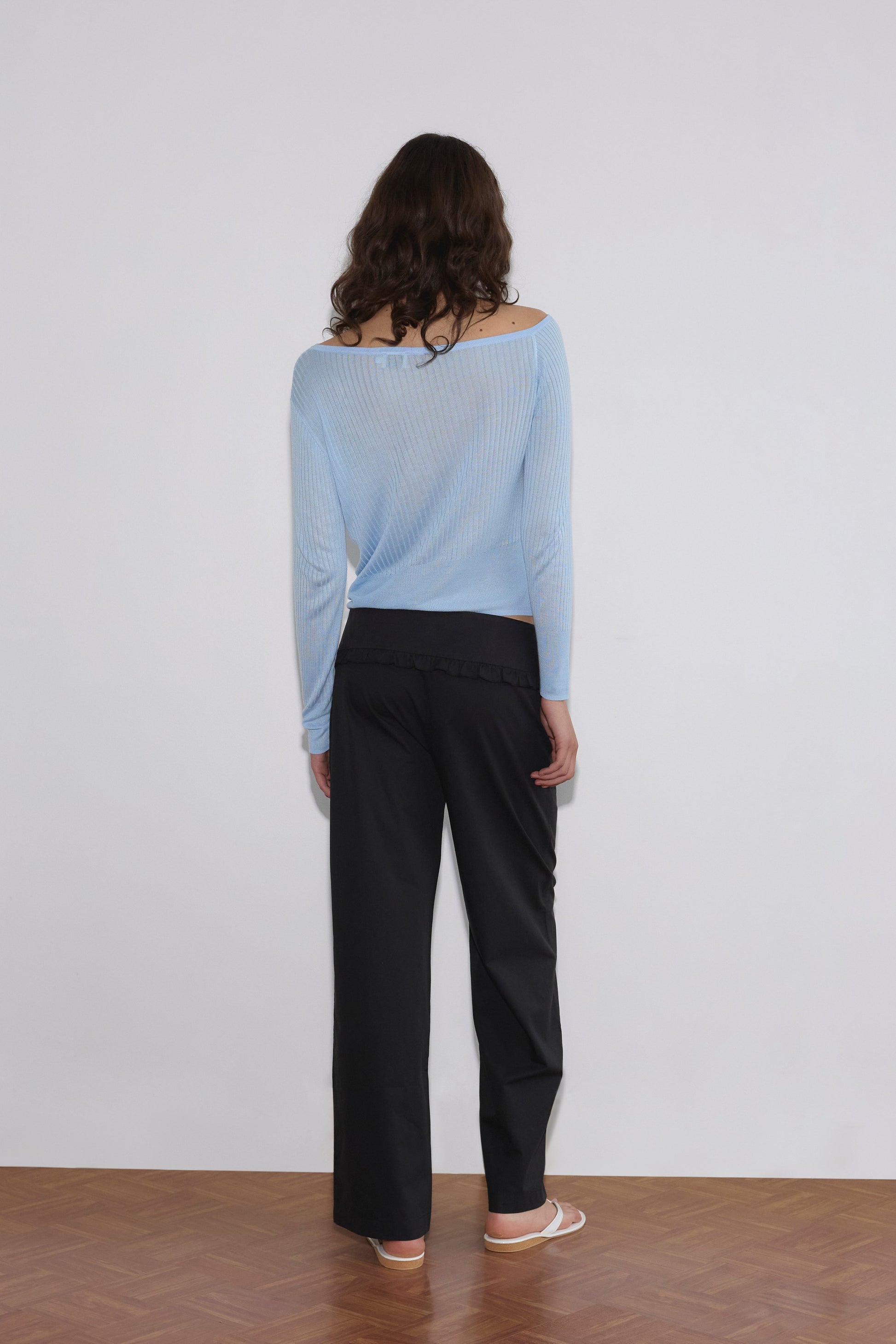 A person with long, wavy hair stands facing a white wall, wearing a sheer light blue Deiji Studios sweater, Flounce Belt Pant in black, and white sandals on a wooden floor.