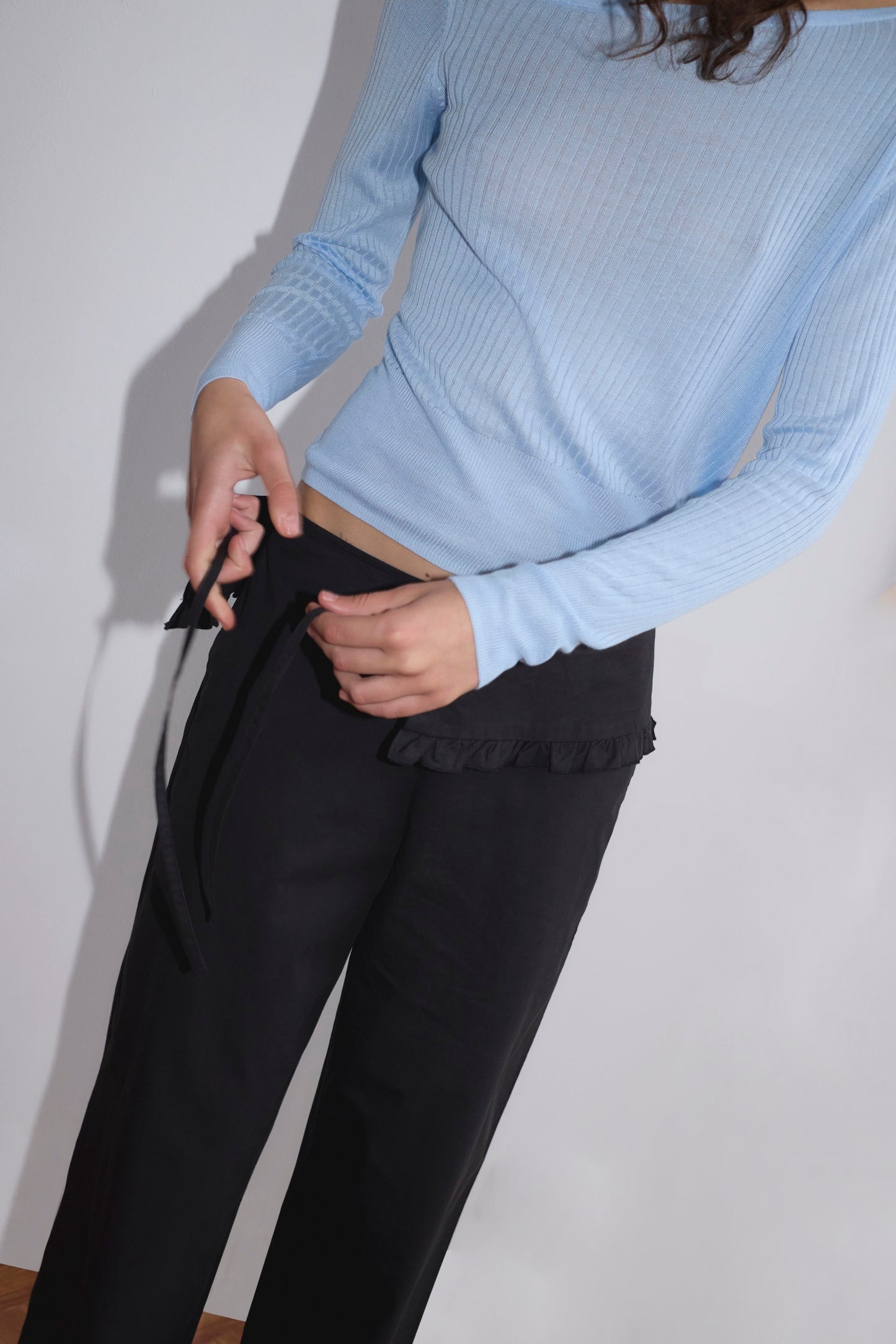 A person, face unseen, wears a light blue ribbed long-sleeve top and the Flounce Belt Pant - Black by Deiji Studios, tying its black belt at the waist. The background is plain and neutral.