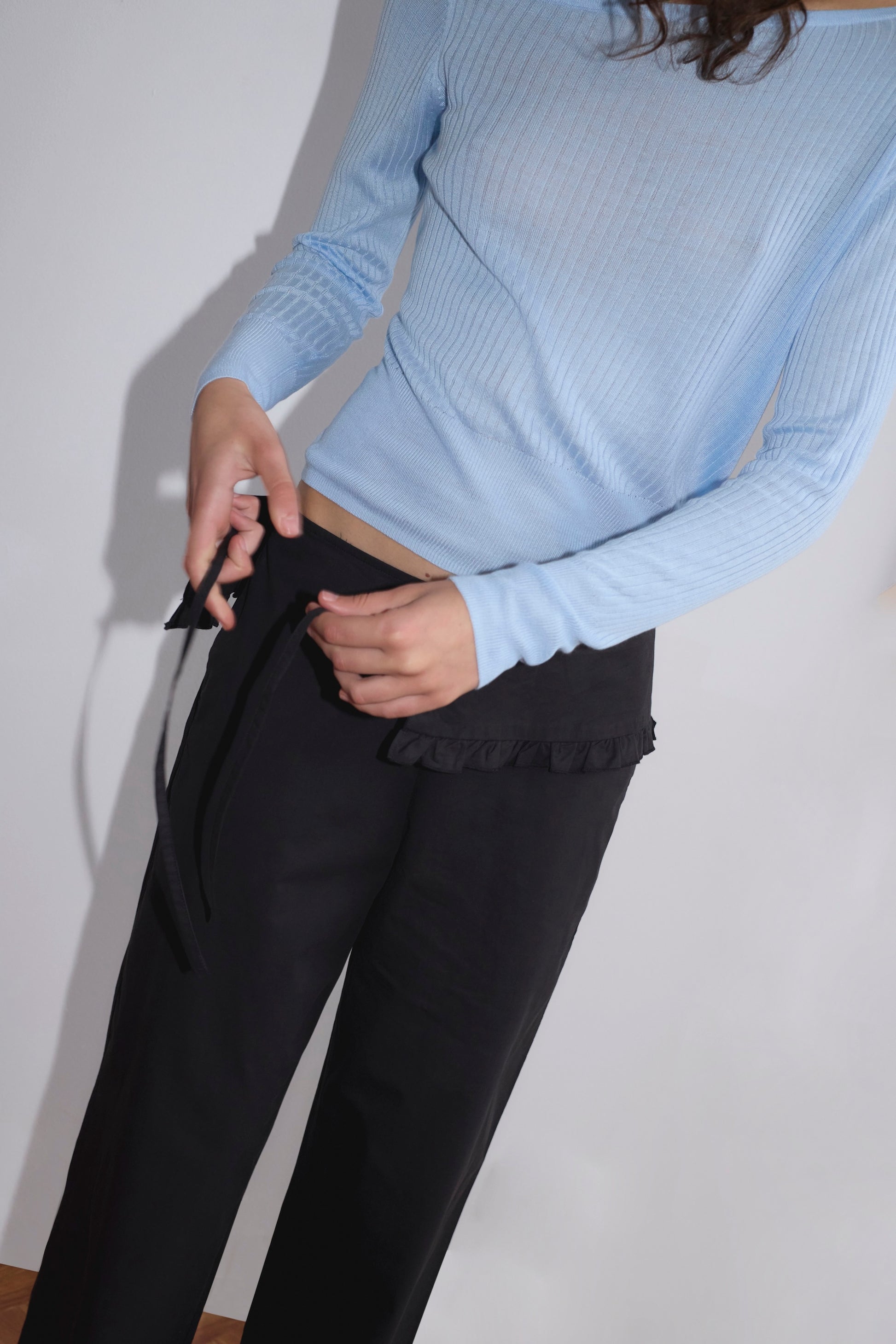 A person, face unseen, wears a light blue ribbed long-sleeve top and the Flounce Belt Pant - Black by Deiji Studios, tying its black belt at the waist. The background is plain and neutral.