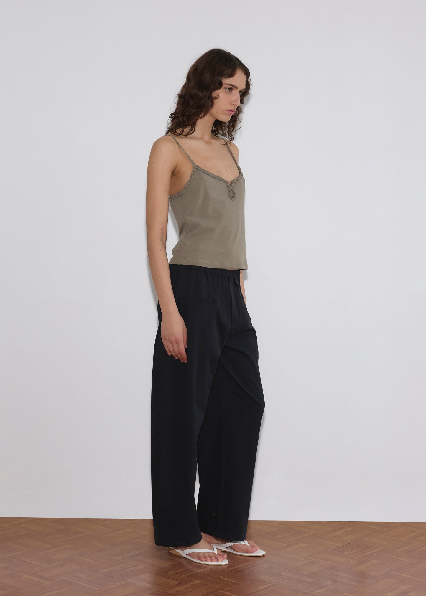 A woman with wavy brown hair wears a beige camisole, Pleat Pant - Black by Deiji Studios, and transparent sandals, standing against a plain white wall on a wooden floor.