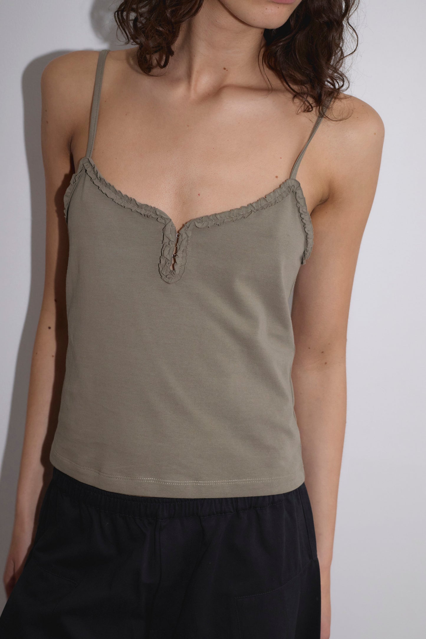 A person with wavy brown hair wears the Clasp Singlet - Khaki by Deiji Studios, featuring thin straps and scalloped trim, paired with black pants, against a white background.