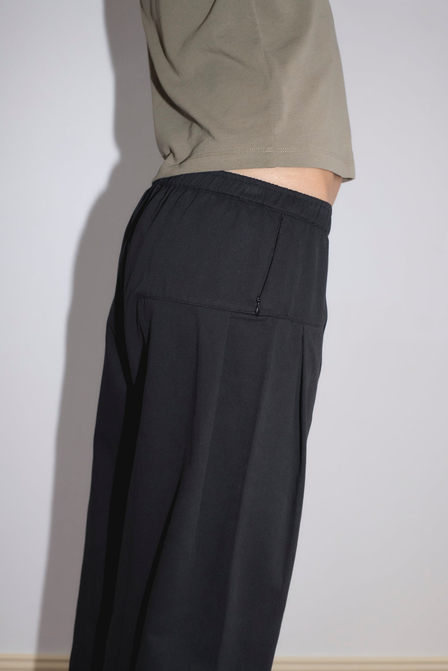 A person stands sideways against a plain light wall, wearing a khaki t-shirt and the Pleat Pant - Black by Deiji Studios, featuring a relaxed straight leg and elastic waistband.