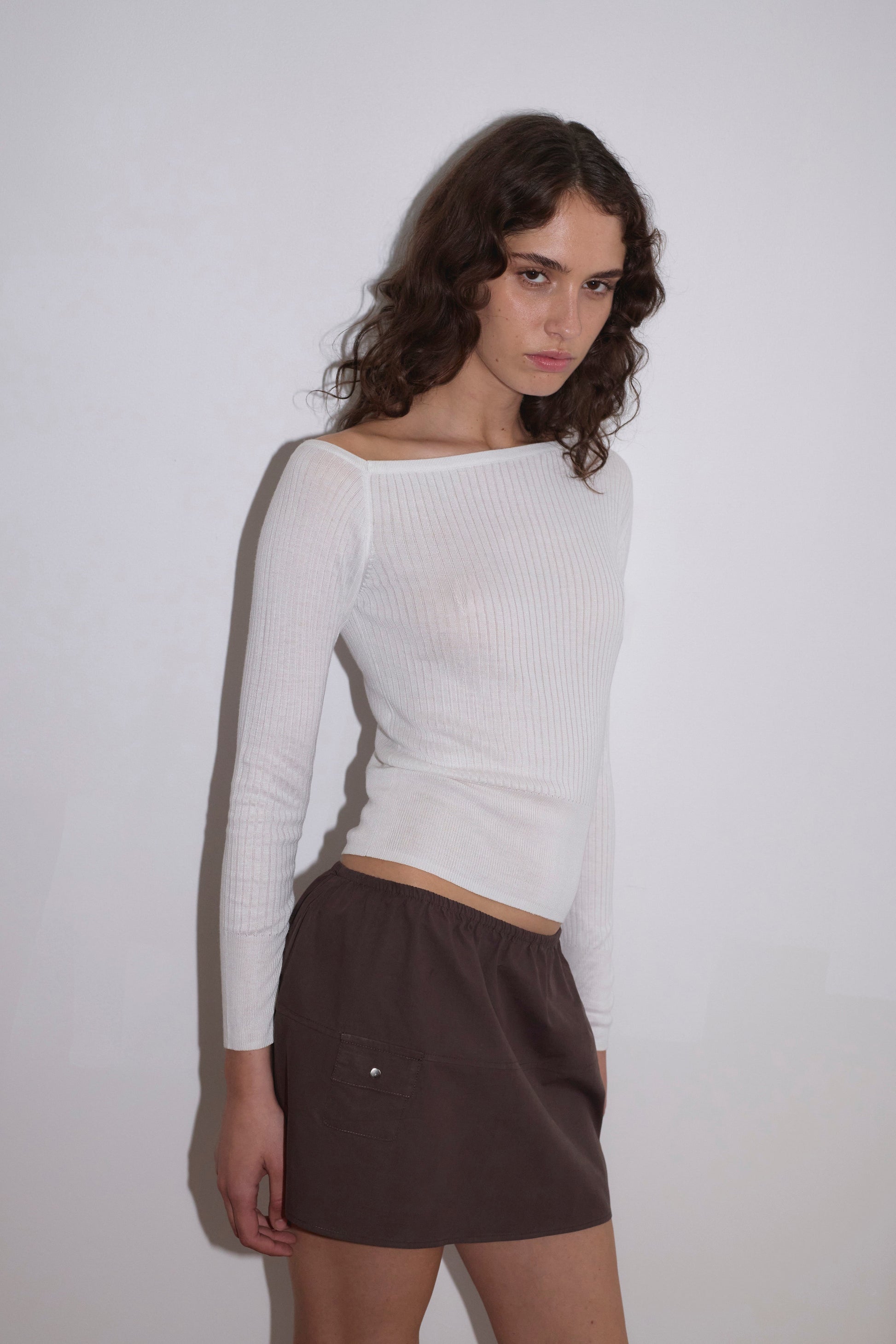 A woman with wavy brown hair wears the Shoulder Knit Top in white by Deiji Studios and a brown mini skirt, standing against a plain white wall and looking slightly to the side.