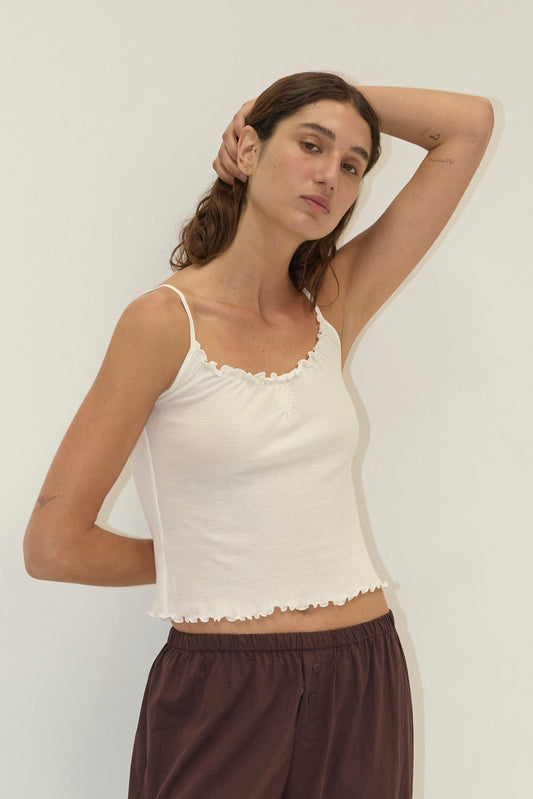 A woman with long brown hair poses against a plain white wall, wearing the Flounce Tank - Cream by Deiji Studios and loose maroon pants. One hand rests behind her head, and she has a neutral expression.
