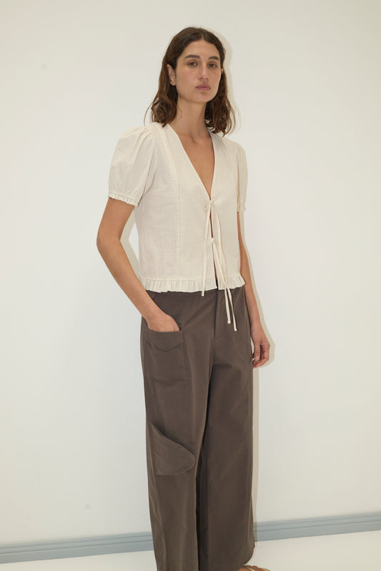 A woman stands against a white wall, wearing the Deiji Studios Pintuck Top - Bone Check in organic cotton with a front tie and loose brown wide-leg pants. She has long, wavy brown hair and one hand in her pocket.
