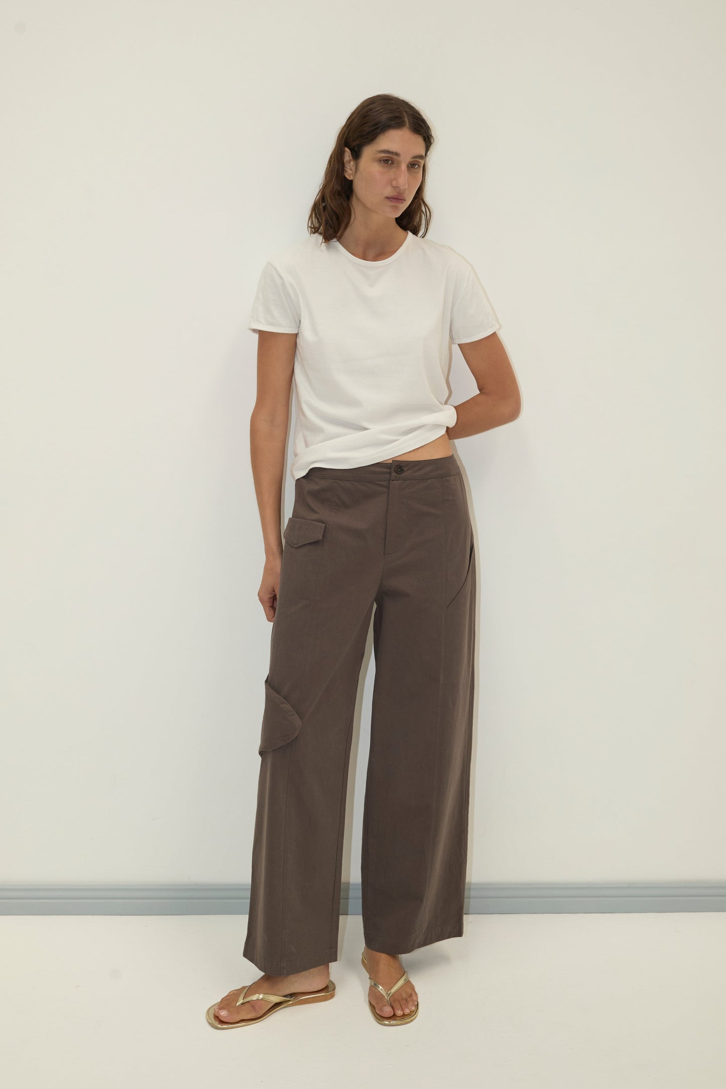 A woman with shoulder-length brown hair and a neutral expression stands against a plain white wall, wearing the Simple Tee - White from Deiji Studios, loose brown trousers with pockets, and brown sandals.