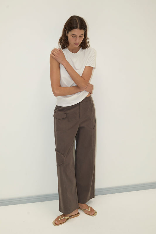 A woman stands against a plain white wall wearing the Pocket Pant in brown with a white t-shirt and flip-flops, her arms crossed over her chest as she looks downward with a neutral expression.