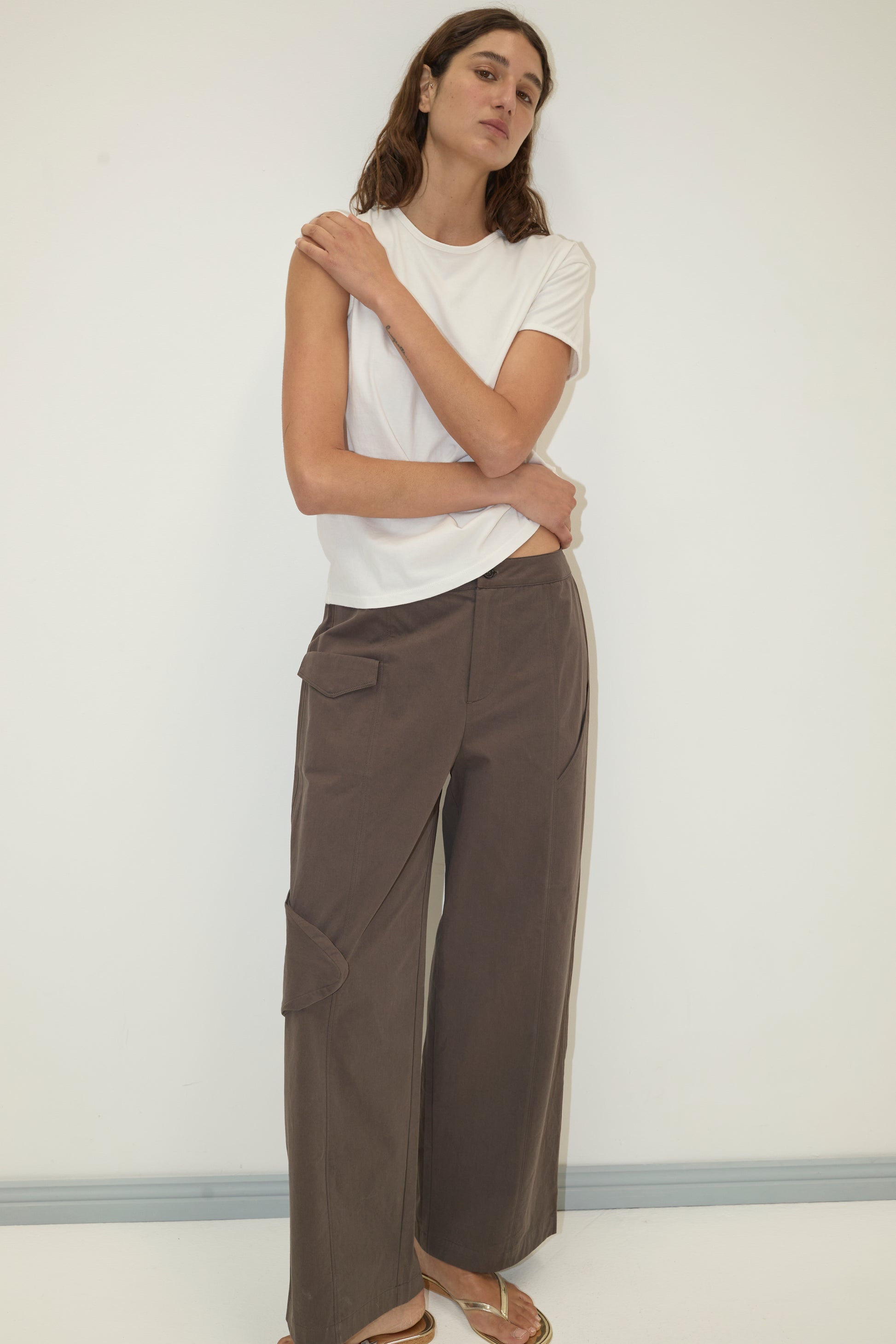 A woman with long brown hair stands against a white wall, arms crossed, wearing Deiji Studios' Simple Tee - White and loose brown pocketed pants, paired with gold sandals for a relaxed, signature look.