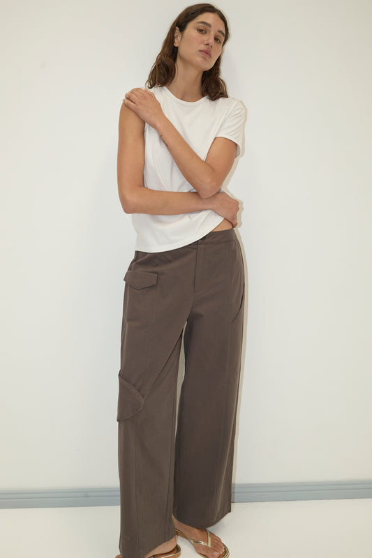 A woman with long brown hair stands against a white wall, arms crossed, wearing Deiji Studios' Simple Tee - White and loose brown pocketed pants, paired with gold sandals for a relaxed, signature look.