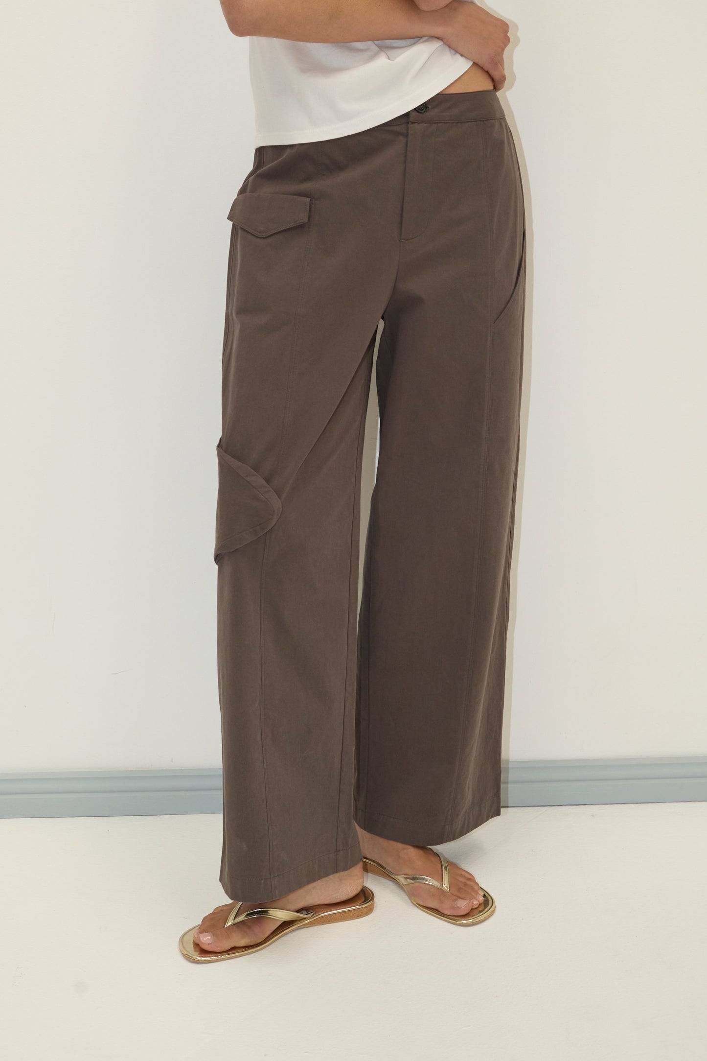 A person wearing the Pocket Pant - Wood, wide-leg brown cargo pants made from organic cotton twill, with a white top and gold sandals stands against a plain light wall. Their face is not visible.