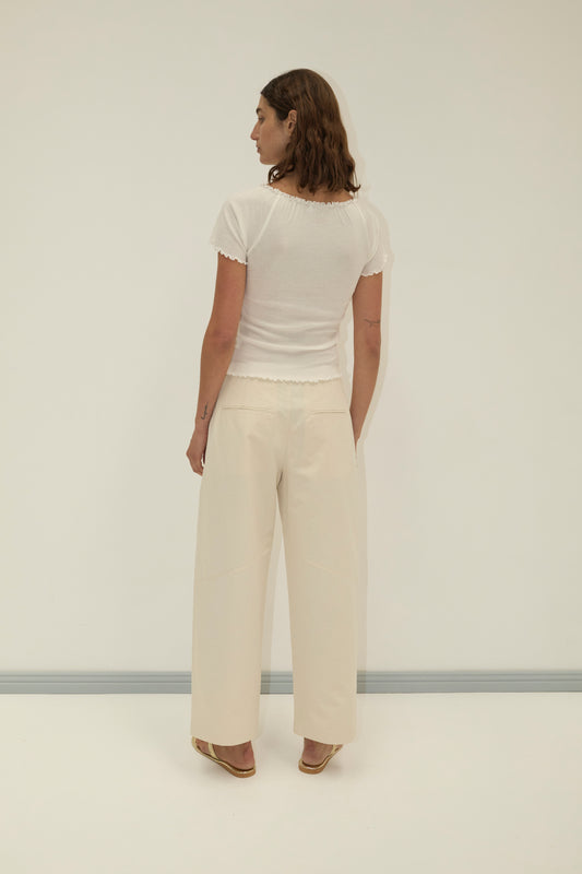 A person with shoulder-length brown hair stands facing away, wearing a short-sleeved white top, Flag Tie Pant in off white, and sandals against a plain light-colored background.