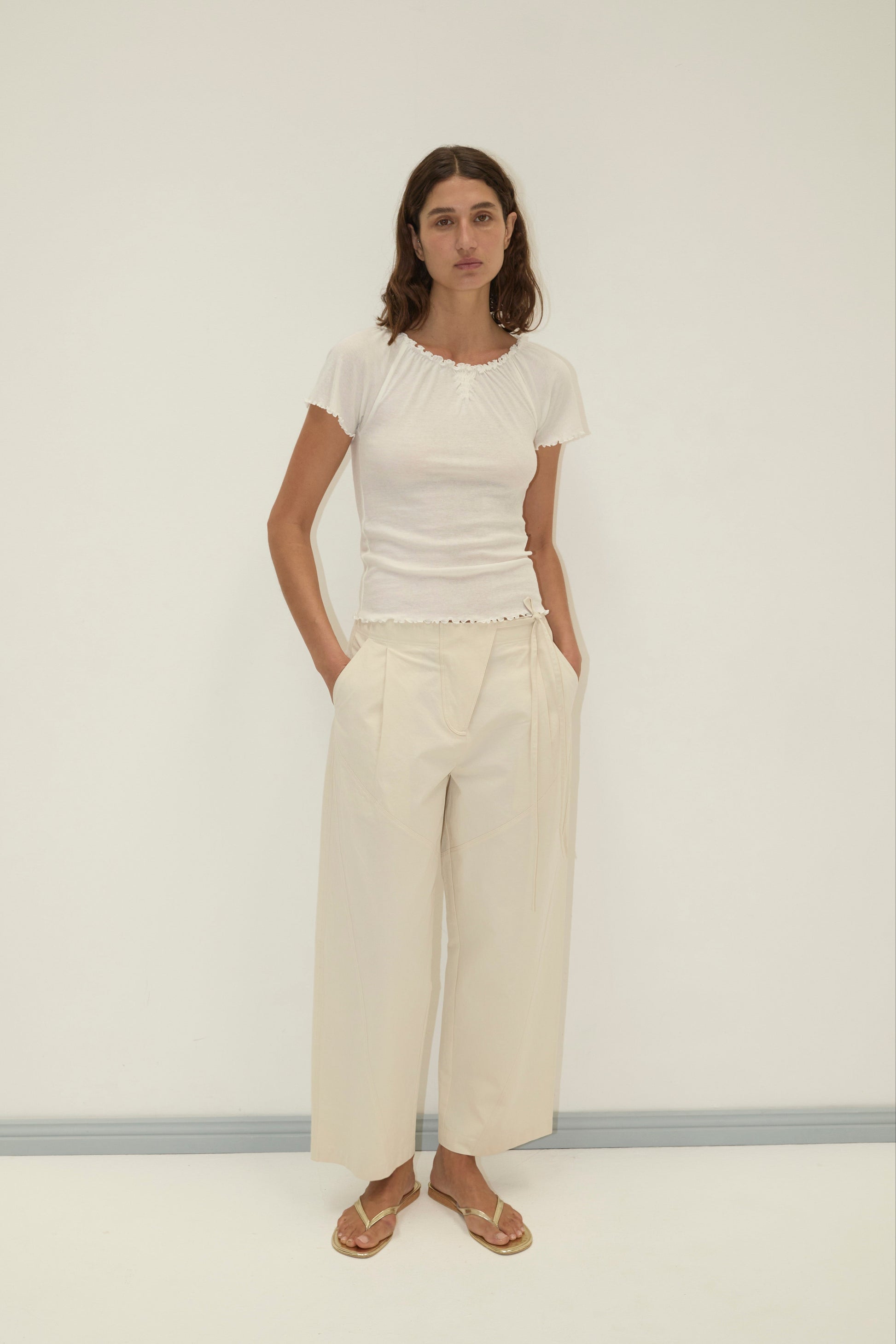 A woman stands against a white wall wearing a fitted white short-sleeve top, Flag Tie Pant in cream, and beige sandals. Her hands are in her pockets and she has a neutral expression.