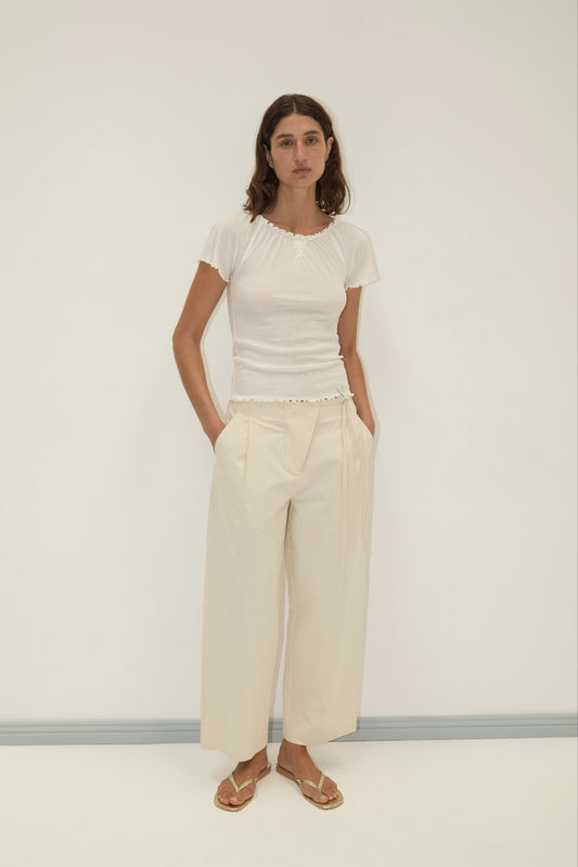 A woman stands against a white wall wearing a fitted white short-sleeve top, Flag Tie Pant in cream, and beige sandals. Her hands are in her pockets and she has a neutral expression.