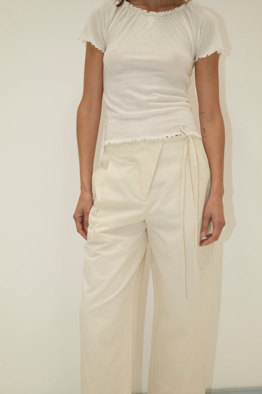 A person models the Flag Tie Pant - Off White, a pair of high-waisted, loose organic cotton pants, with a fitted white short-sleeve top against a plain light background. Their face is not visible.